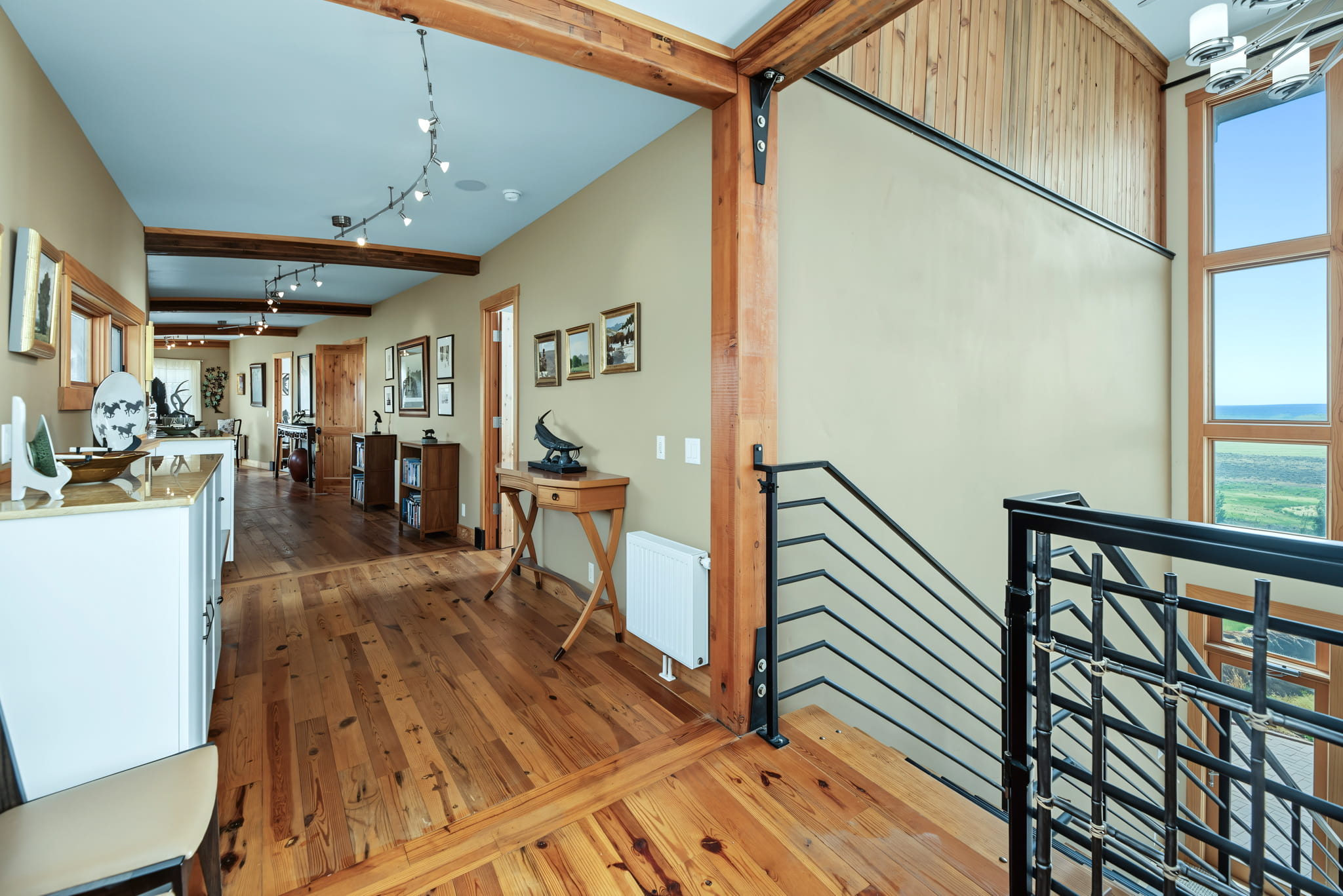 Hallway with wooden floor and exposed beams, decorated with artwork and sculptures, leading to multiple doors. Large windows by the staircase provide a scenic view of greenery outside.