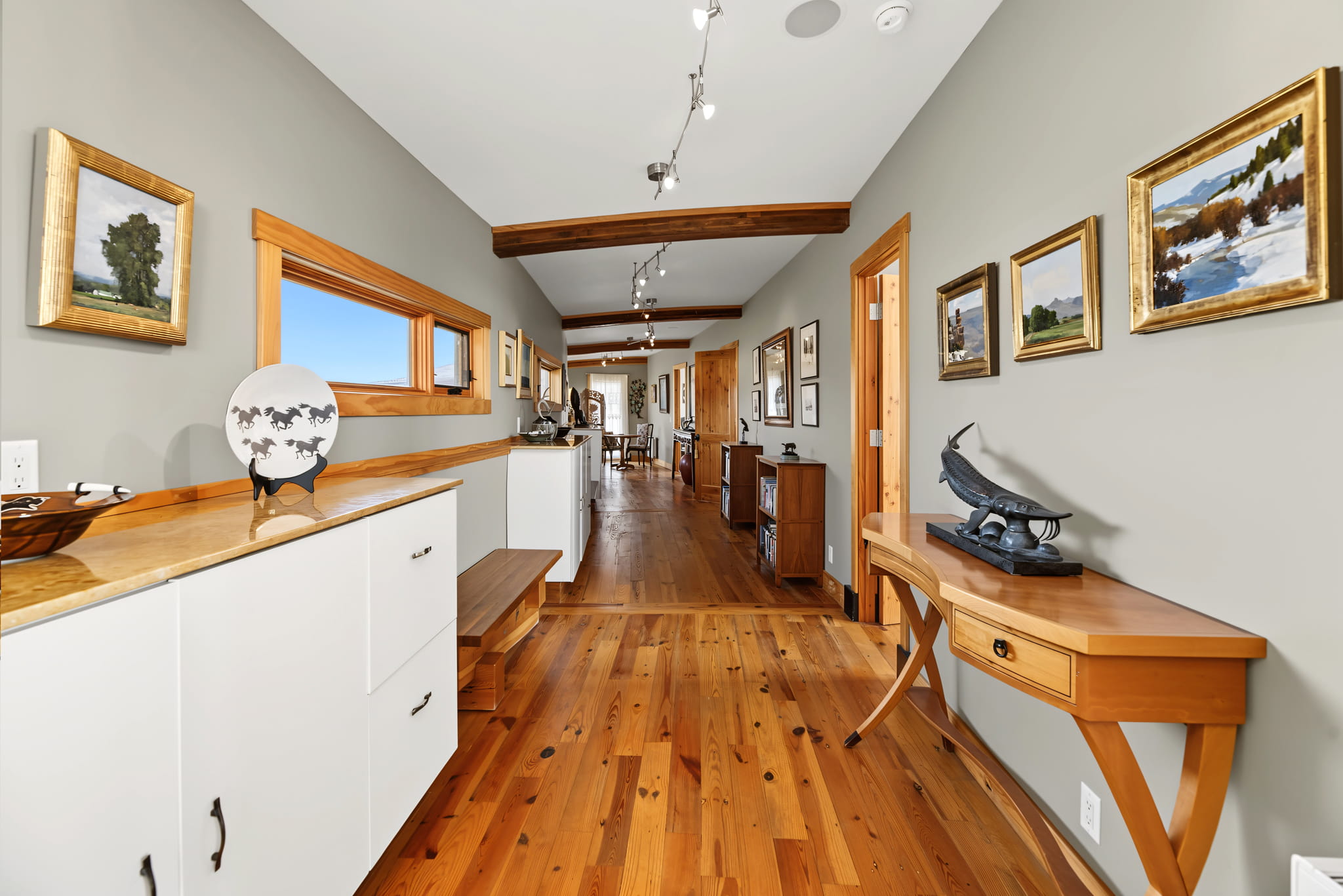 A hallway with light wood floors, white cabinets, and beige walls adorned with landscape paintings. A wooden table with a metal fish sculpture sits on the right. The space is bright and modern.