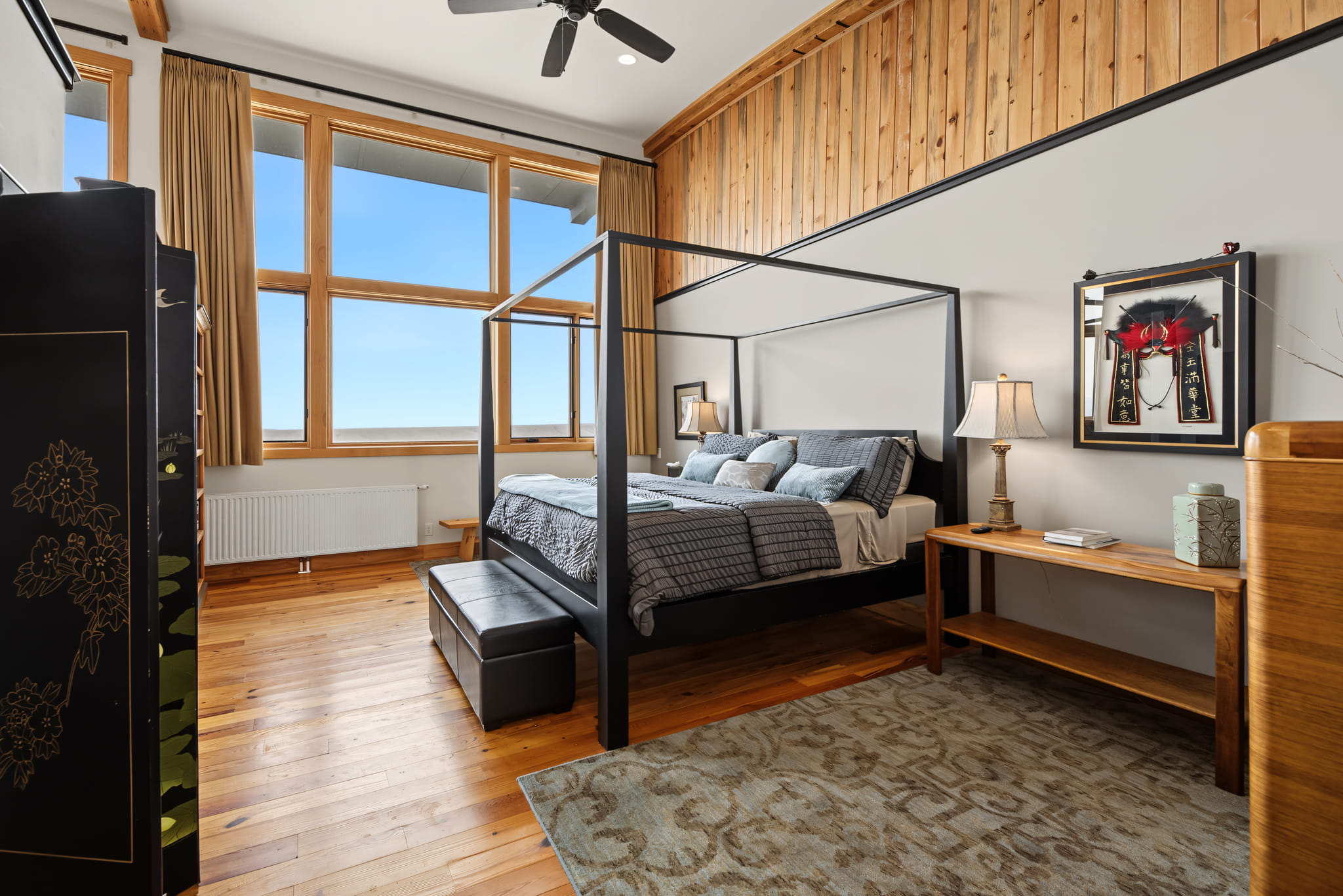 A modern bedroom with a black four-poster bed, large windows, wood floors, and wood paneling on the upper wall. There is a rug, nightstands with lamps, and framed art above one nightstand.