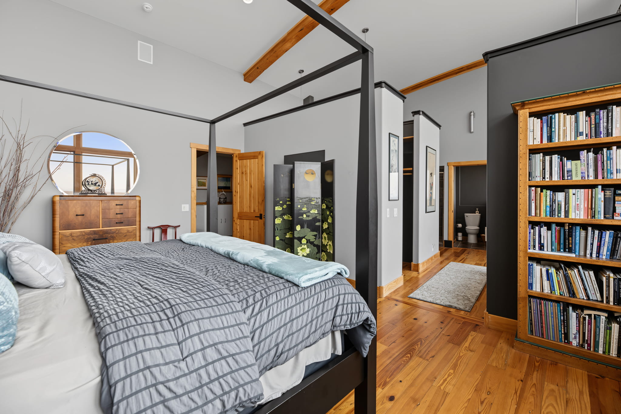 A cozy bedroom with a four-poster bed, gray bedding, wooden floors, a bookshelf, dresser with a round mirror, and a partial view of a bathroom down the hallway.