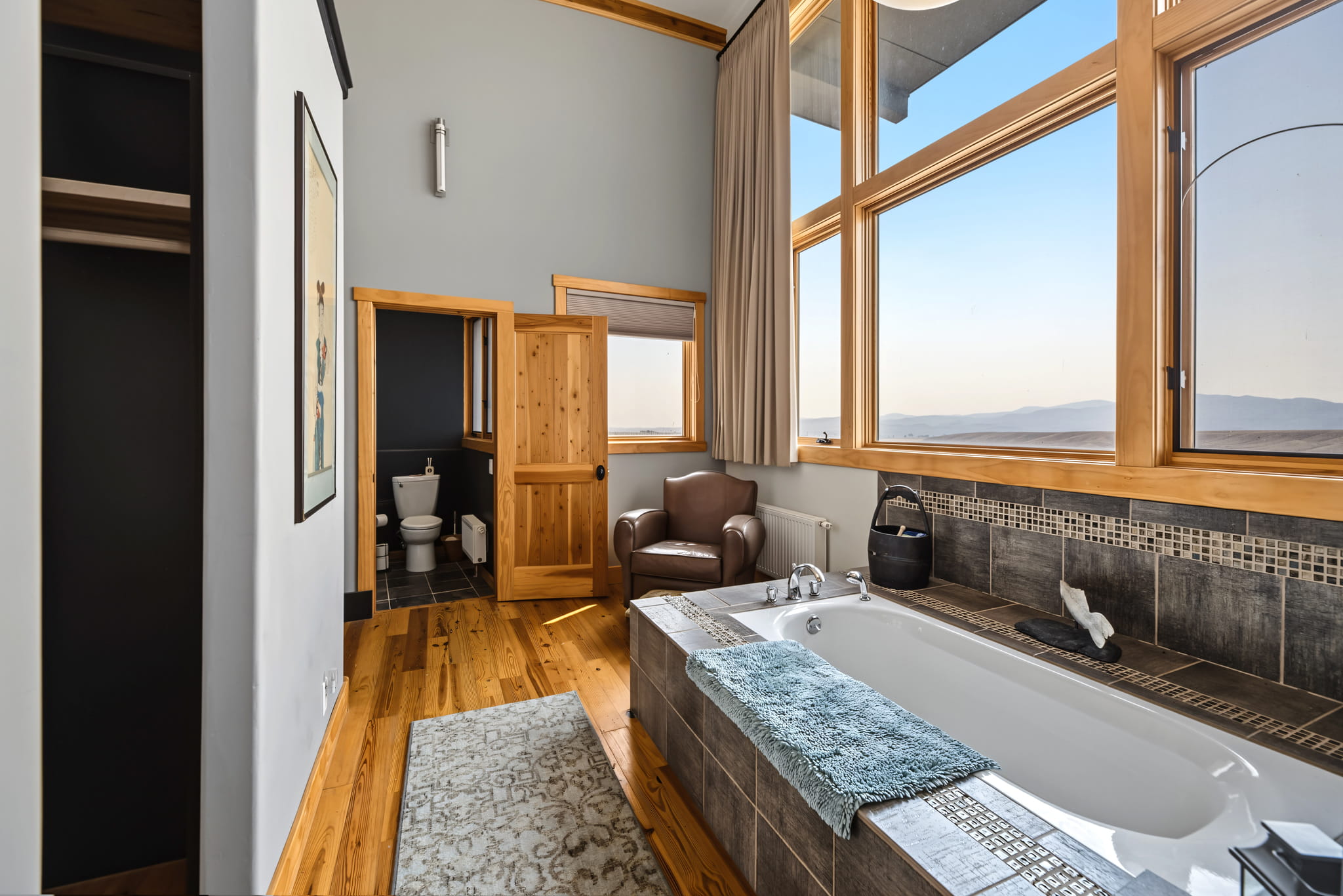 Modern bathroom with a large bathtub, towel, and armchair by big windows showing a scenic view. Wooden floors, open closet, rug, and a door leading to a toilet are also visible.