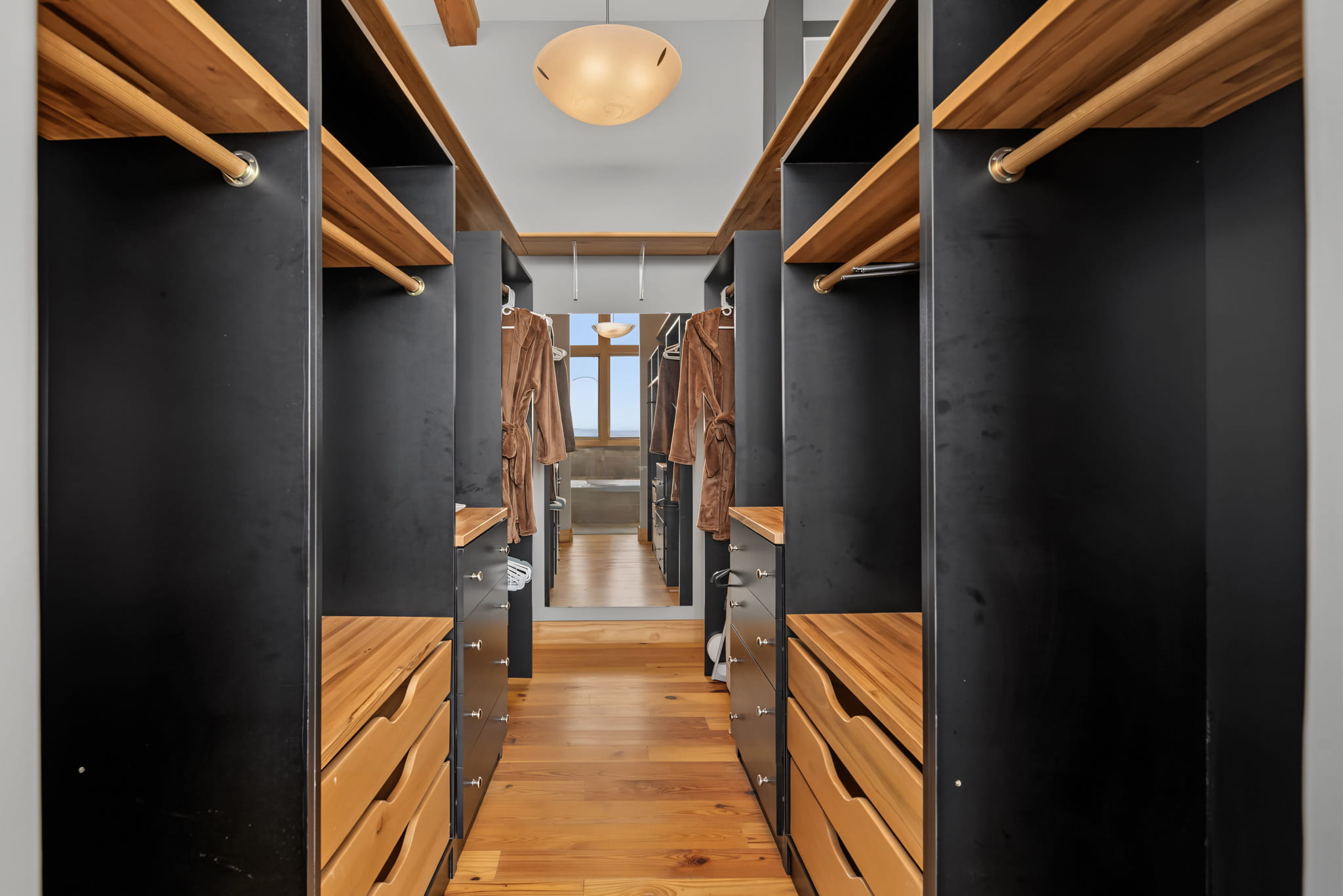 A modern walk-in closet with wooden shelves, black panels, drawers, and hanging rods, featuring a full-length mirror at the end and a light fixture on the ceiling.