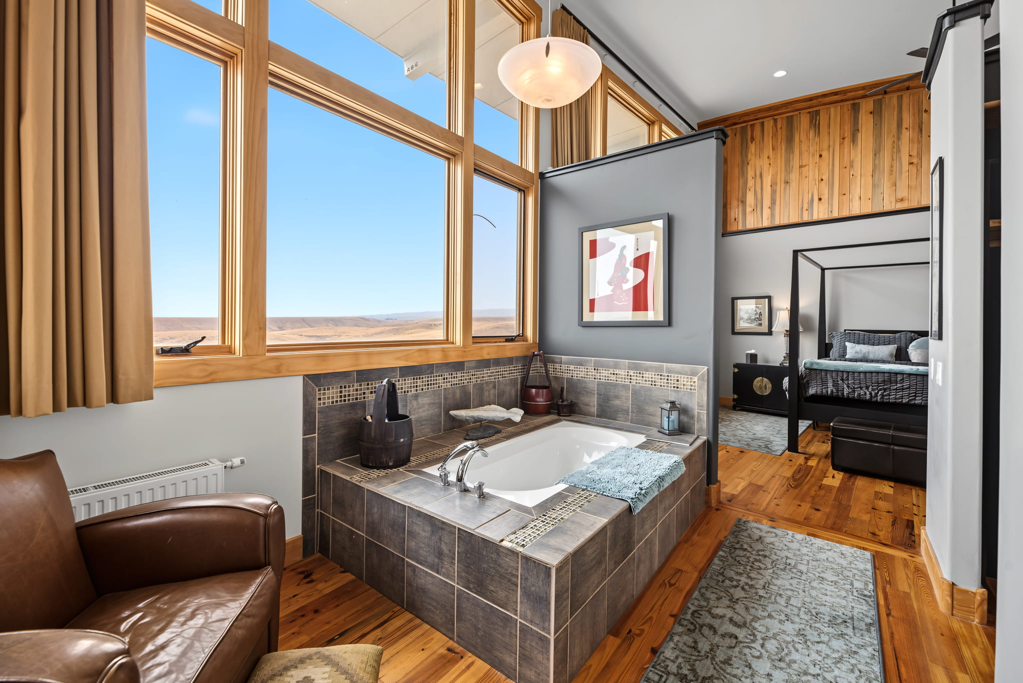 A modern bedroom with an en-suite tub surrounded by tile, large windows with a scenic view, wooden floors, a leather chair, and a cozy bed in the background.