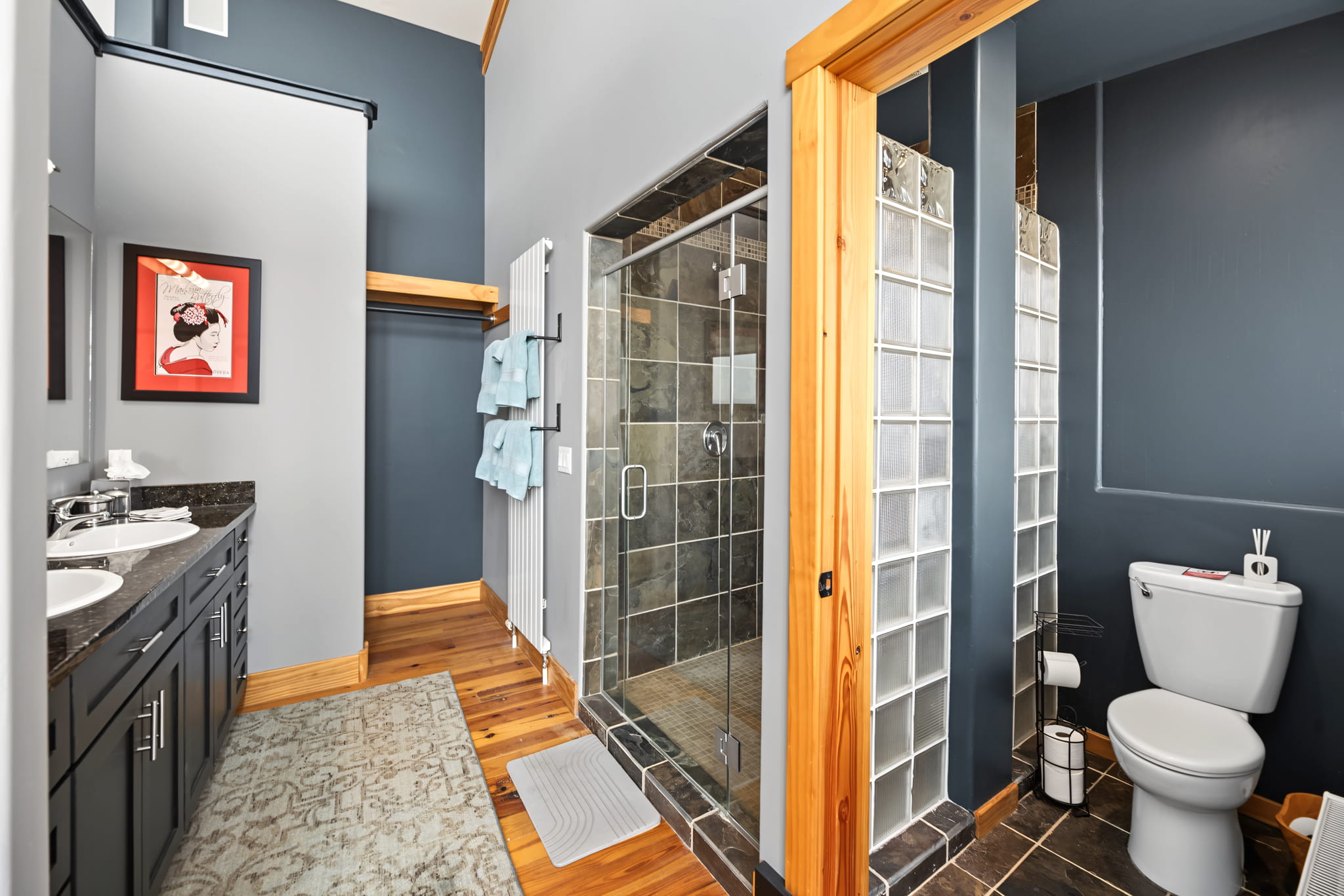 Modern bathroom with double sinks, a glass-enclosed shower, a toilet, dark cabinets, wood trim, blue-gray walls, and art above the towel rack.