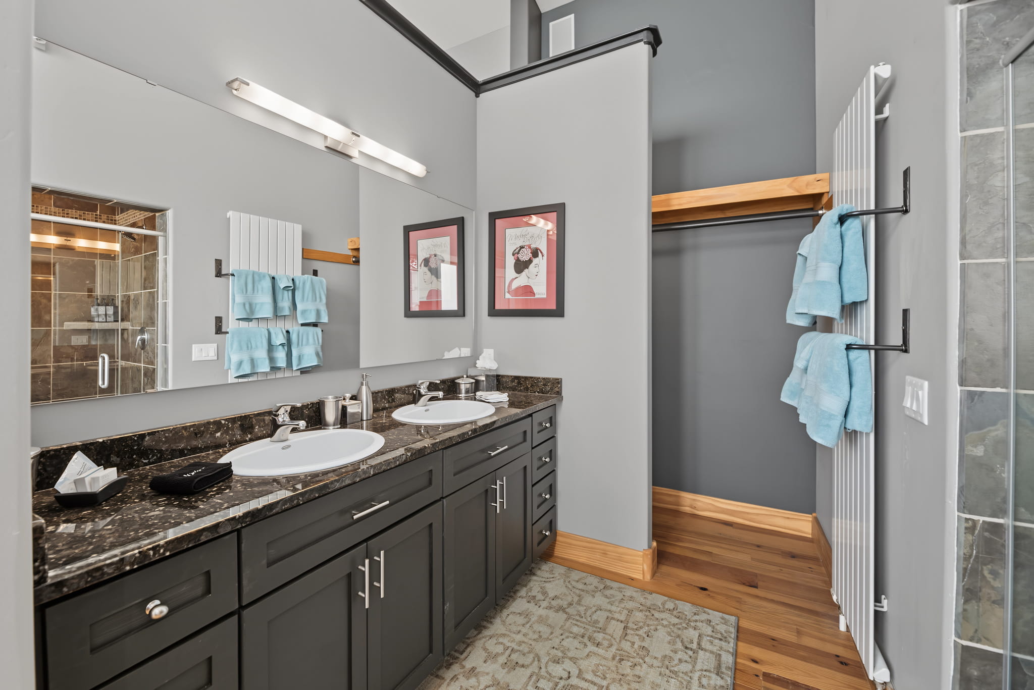 Modern bathroom with double sinks, dark cabinets, granite countertop, two framed art prints above the vanity, blue towels on a rack, a large mirror, and a walk-in closet with wooden shelves and rod.