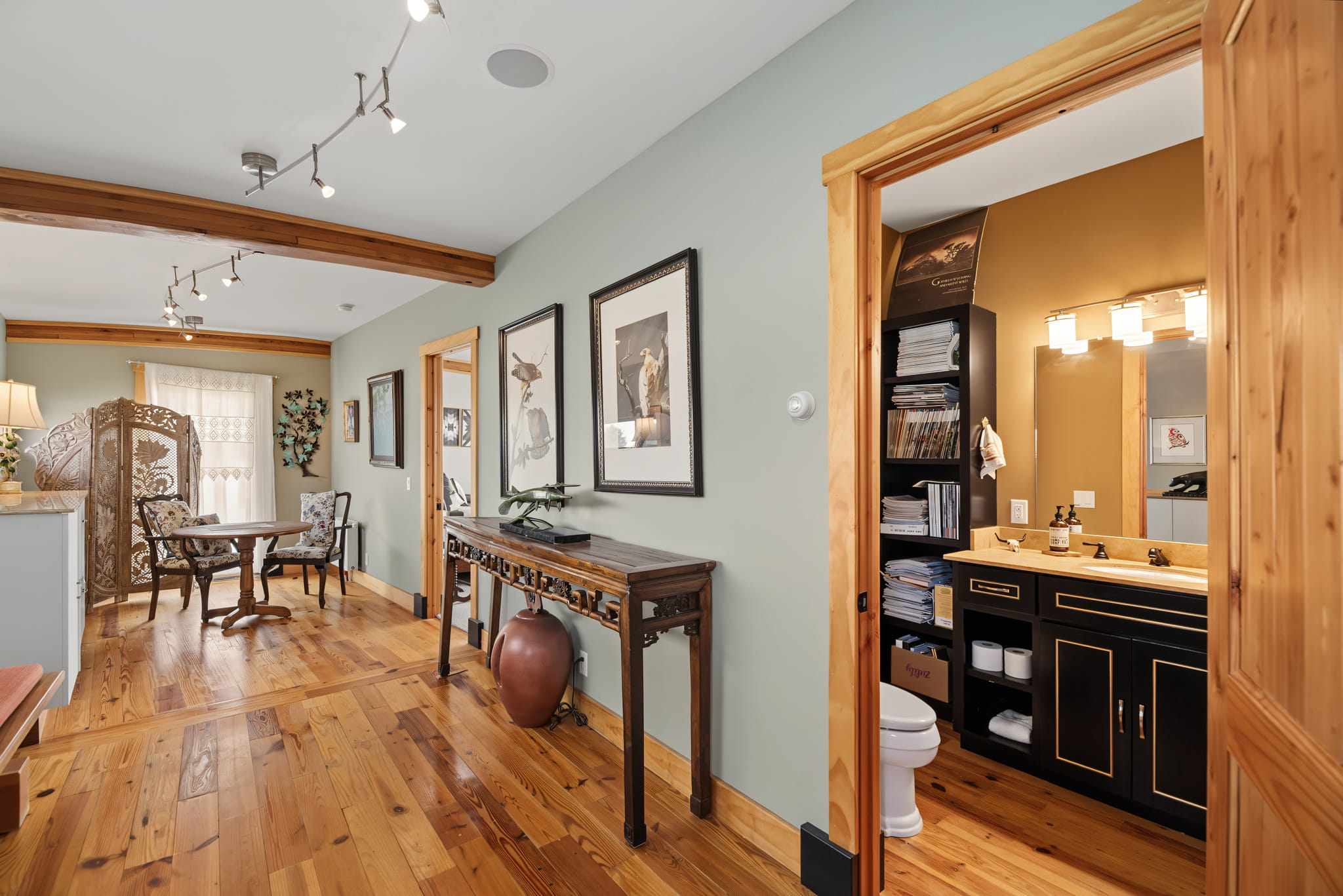 Hallway with wood floors, Asian-inspired decor, and framed art. Theres a bathroom with black vanity to the right, and a dining area with a round table and chairs in the background. Exposed wooden beams accent the ceiling.