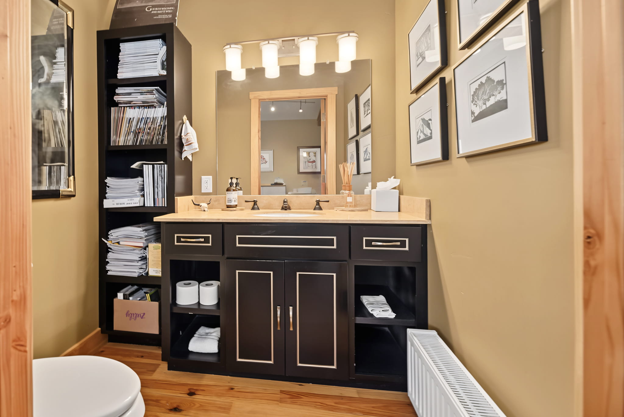 Bathroom with a black vanity, double sink, large mirror, shelves stacked with neatly folded towels and magazines, framed pictures on tan walls, and a wooden floor.