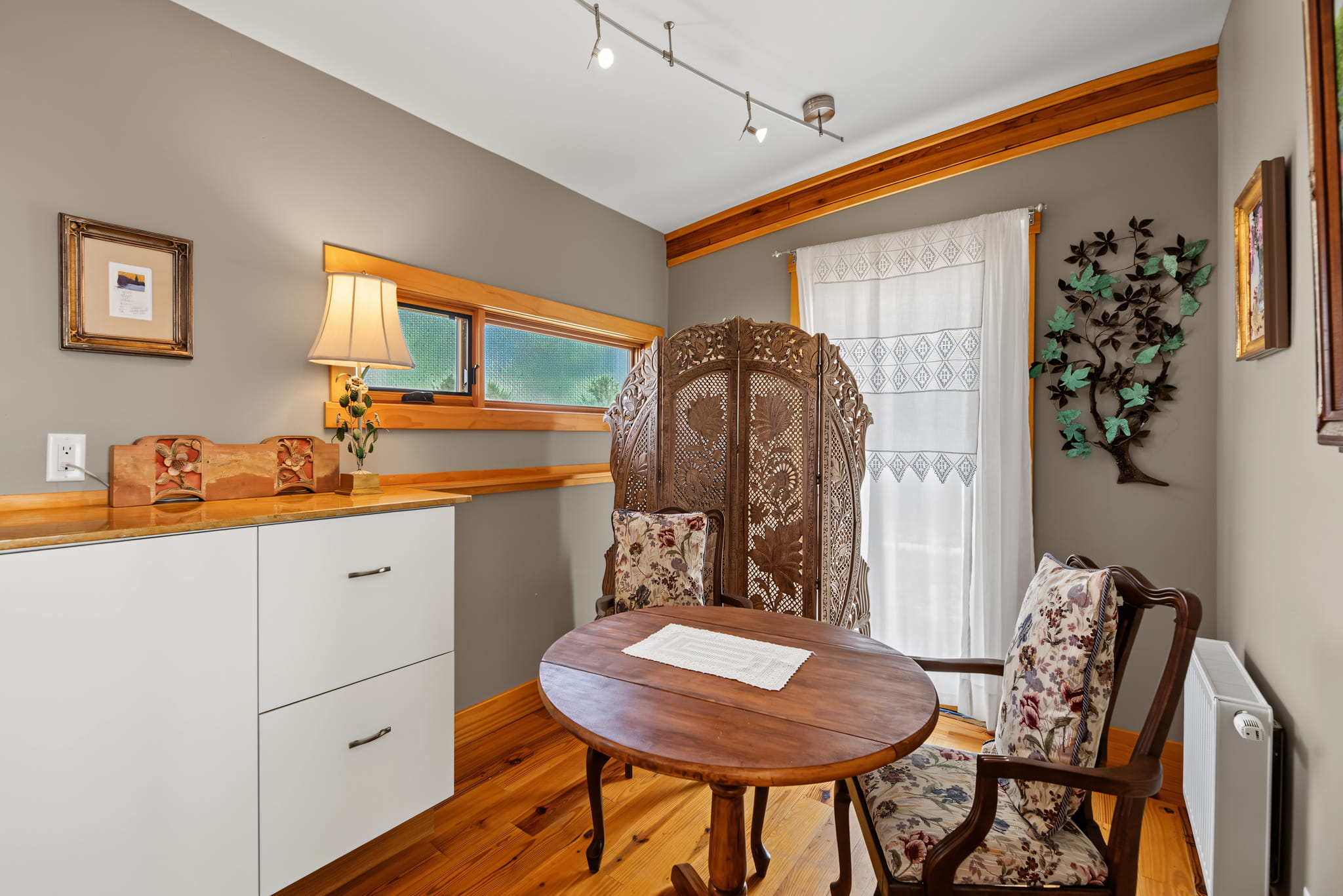 Cozy room with a round wooden table, two floral chairs, a folding privacy screen, a wall-mounted lamp, white cabinets, art on the walls, and a window with sheer curtains.