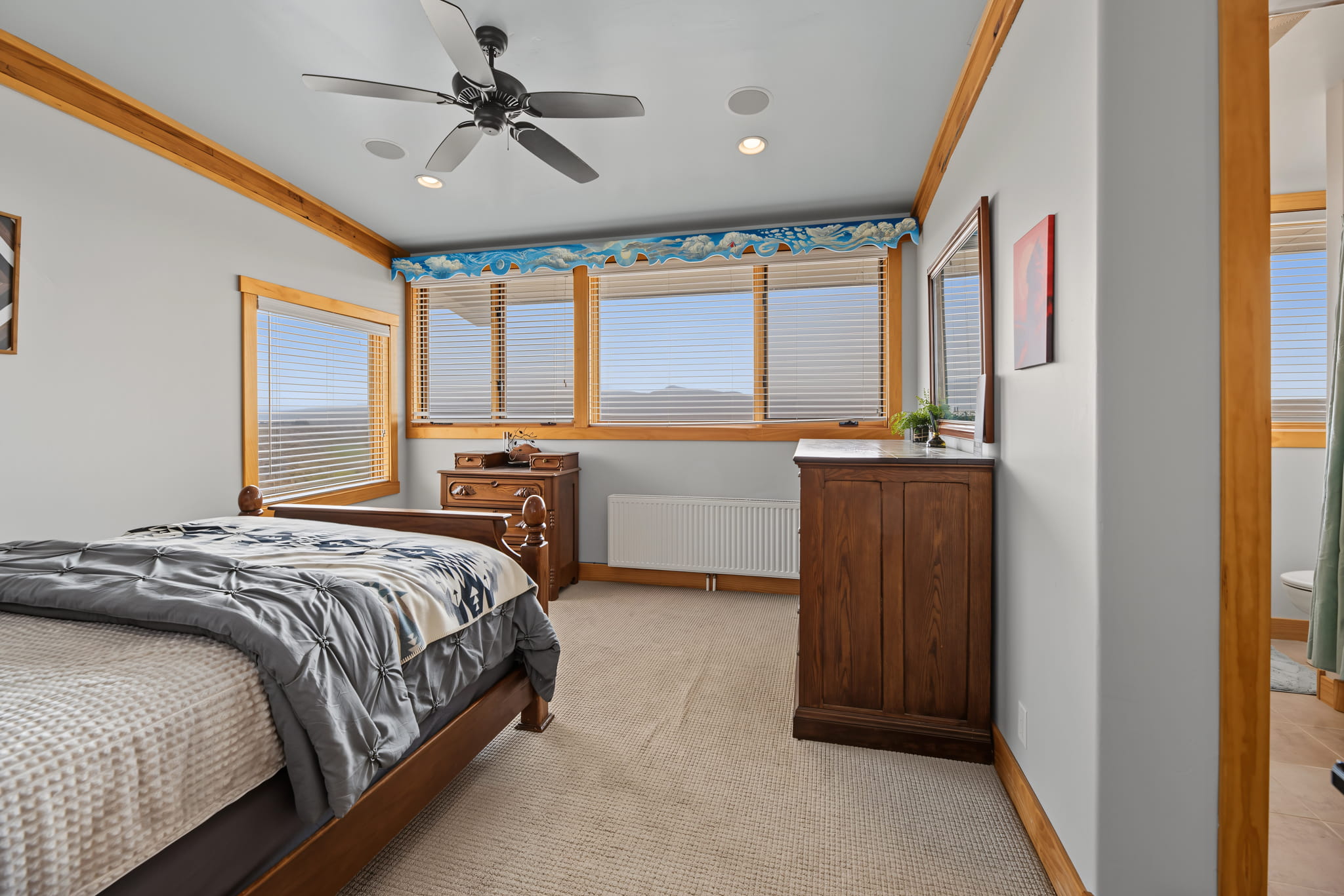 A cozy bedroom with a bed, wooden furniture, a ceiling fan, and large windows letting in natural light. Blue and white wave-themed trim decorates the tops of the windows.