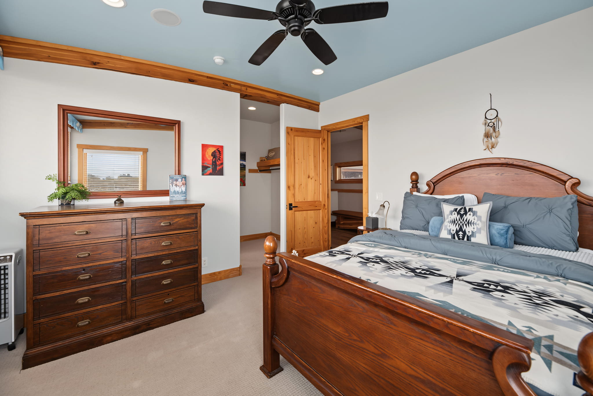 A cozy bedroom with a wooden bed, blue-gray bedding, a wooden dresser with a mirror, a ceiling fan, and wooden trim. A dreamcatcher hangs on the wall, and a small plant sits on the dresser.
