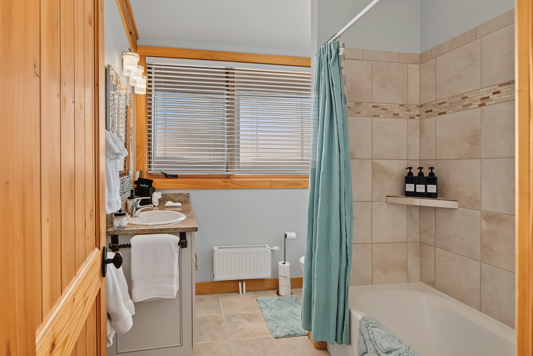 A bathroom with a wooden door, a sink with towels, a toilet, a bathtub with a teal shower curtain, and bottles on a shelf. Large window with blinds, beige tiled walls, and a bath mat on the tiled floor.