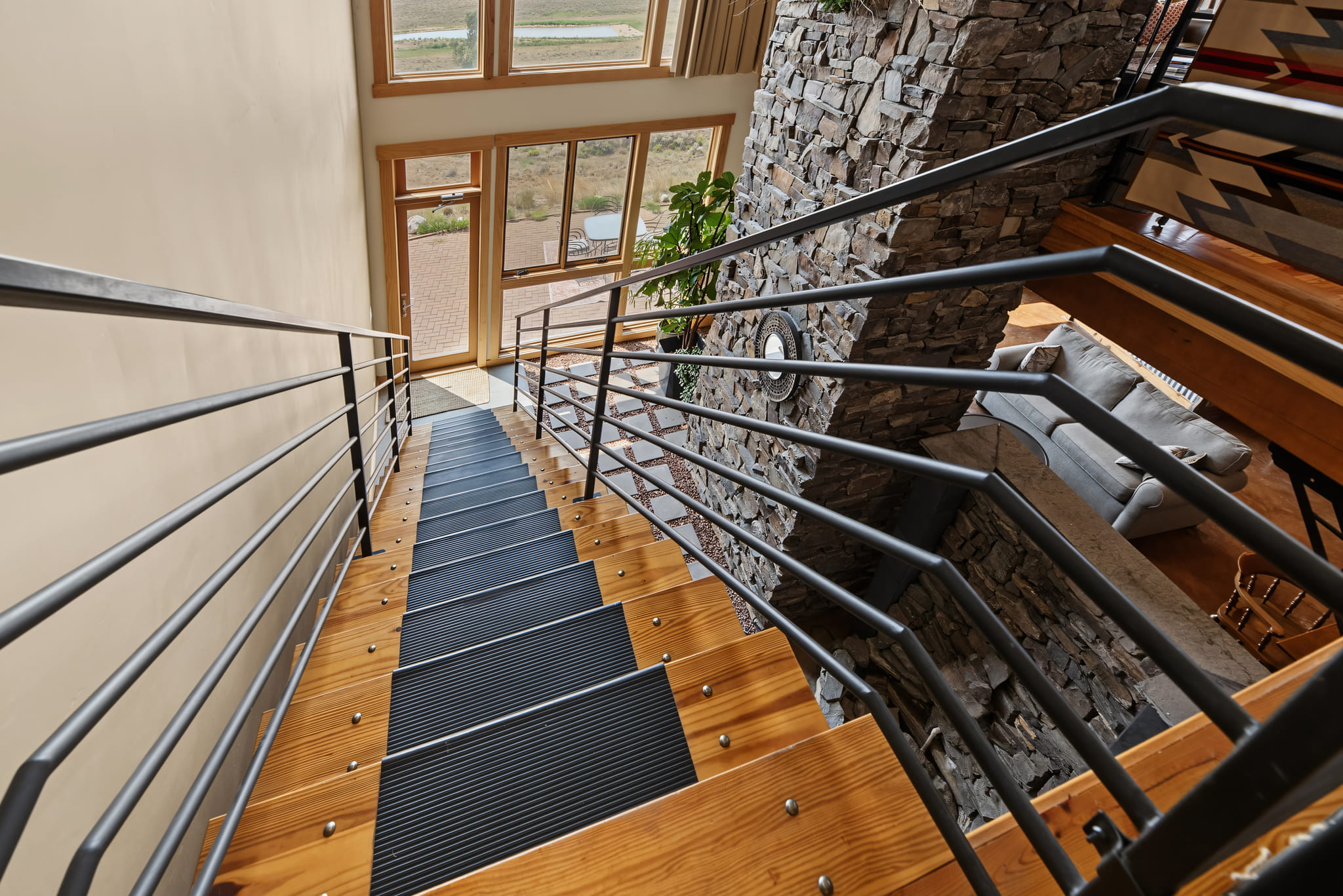 View from the top of a modern wooden staircase with metal railings, looking down toward a large window and stone wall in a bright living area. A sofa and indoor plant are visible near the bottom.