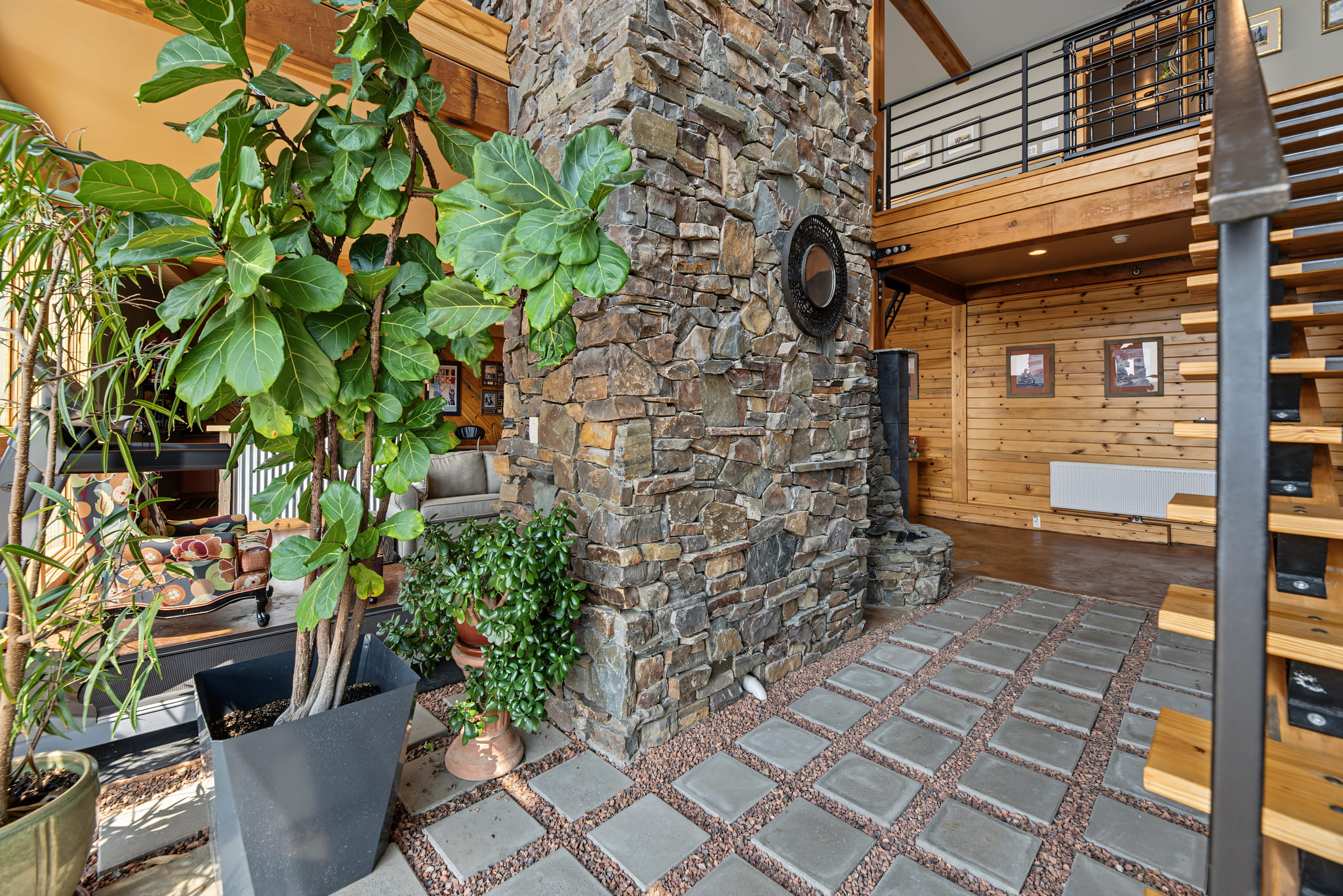 Spacious interior with stone fireplace, wooden walls, large green houseplants, modern staircase, and sunlight streaming in from the left. Cozy and rustic decor throughout.