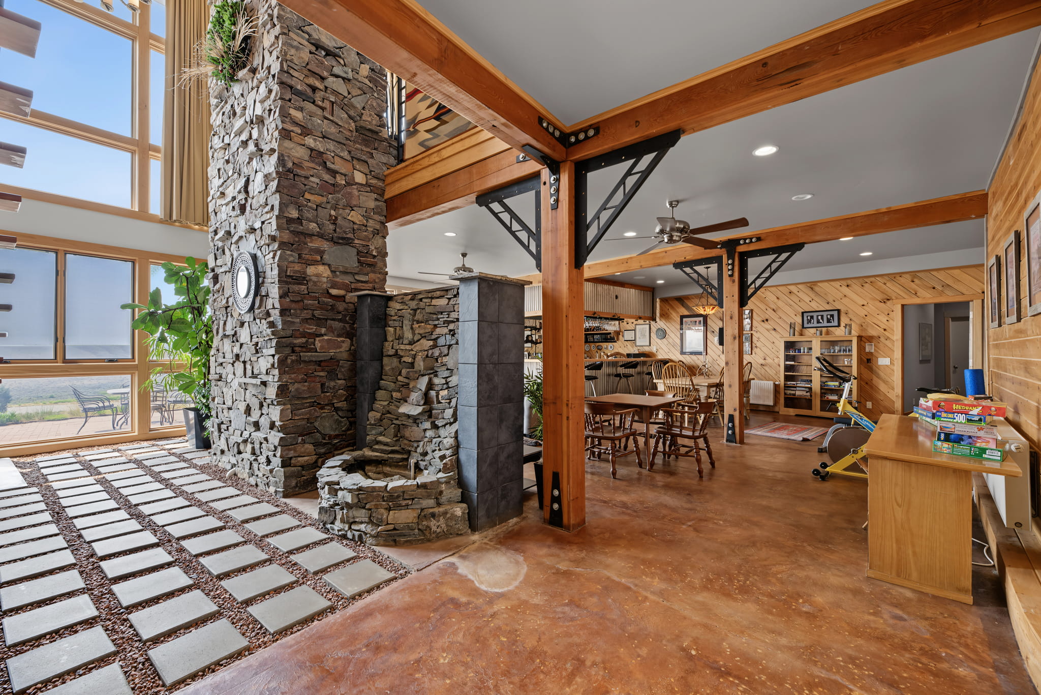 Spacious open-concept room with stone fireplace, exposed beams, and wood accents. Dining area with wooden table and chairs, play area, large windows, and natural light create a warm, inviting atmosphere.