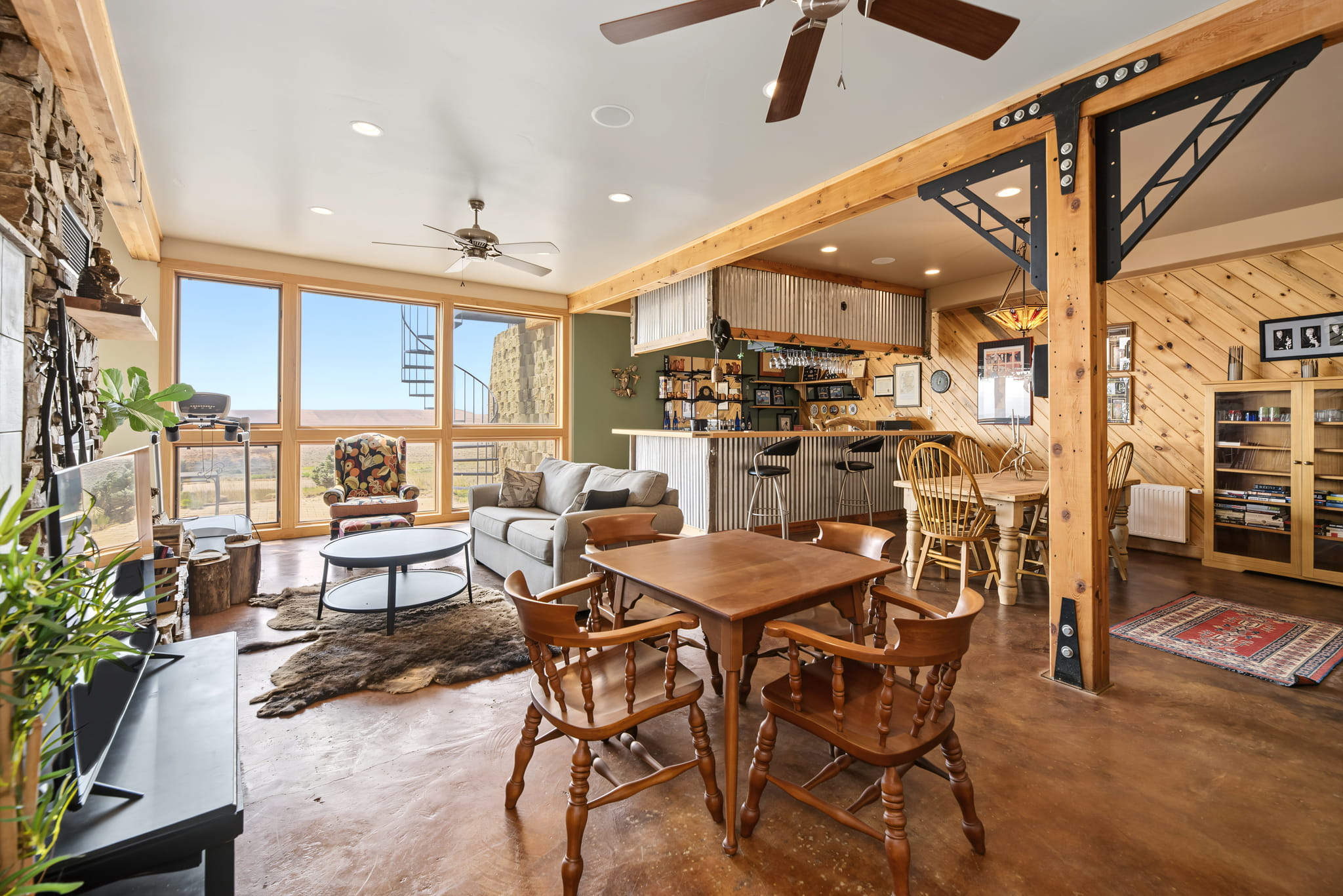 Open-concept living room with wood and metal accents, cozy seating area, small dining table, bar, and large windows offering a scenic view. Spiral staircase visible in the corner. Warm, inviting decor.