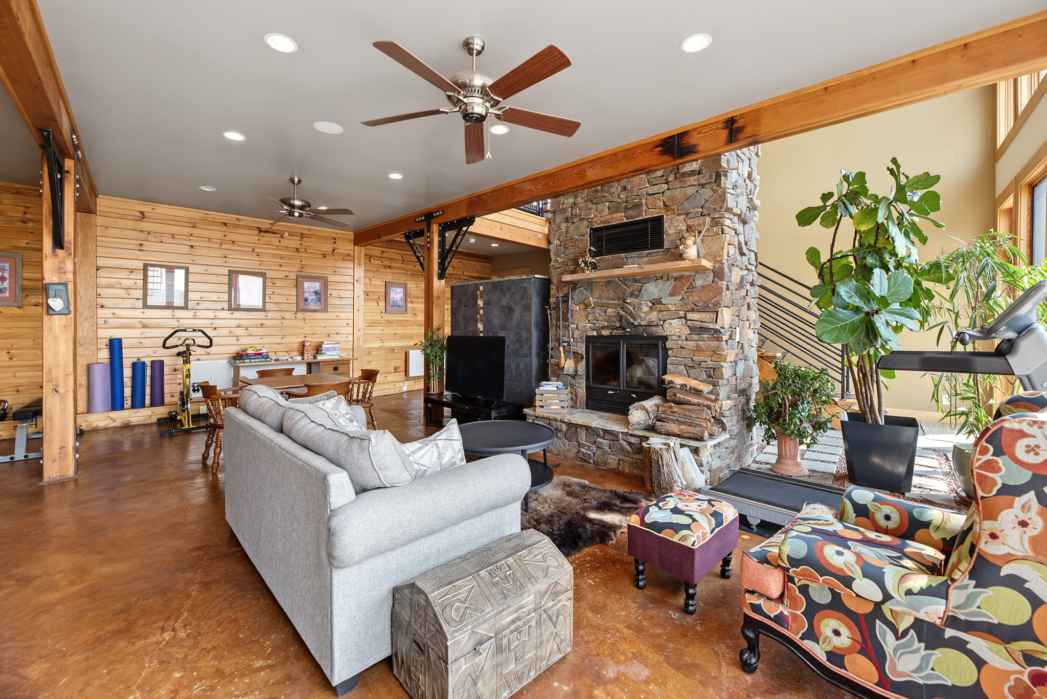 Spacious living room with wood walls, stone fireplace, gray sectional sofa, colorful armchair, plants, ceiling fans, and desk area, featuring large windows and warm, natural light.
