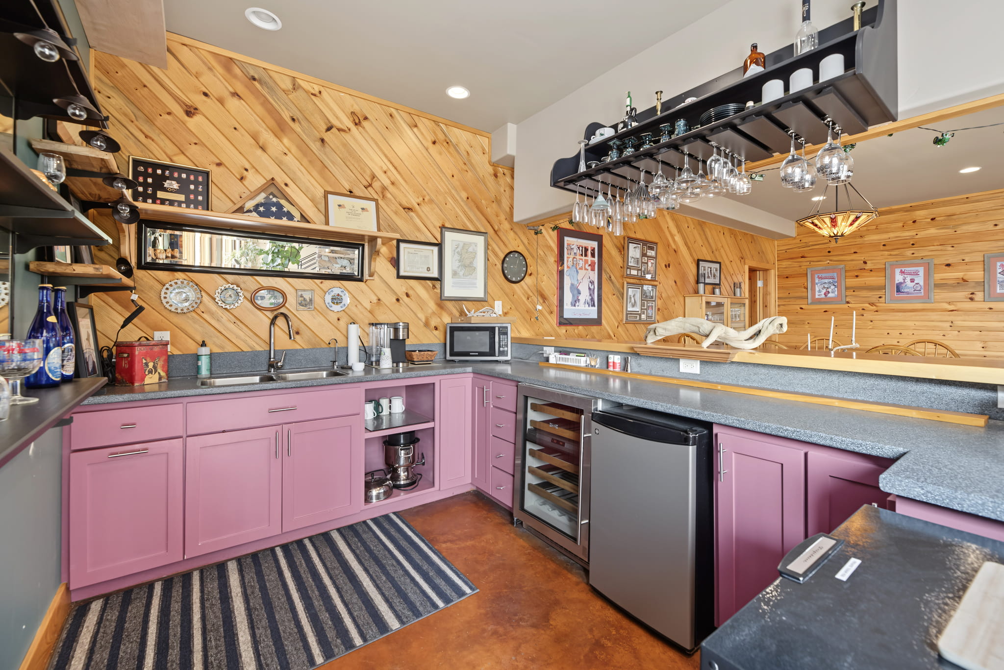 Home bar with purple cabinets, wine fridge, microwave, glass rack, and open shelving. Wood-paneled walls feature framed art, plates, and certificates. Modern lighting and a striped rug complete the space.