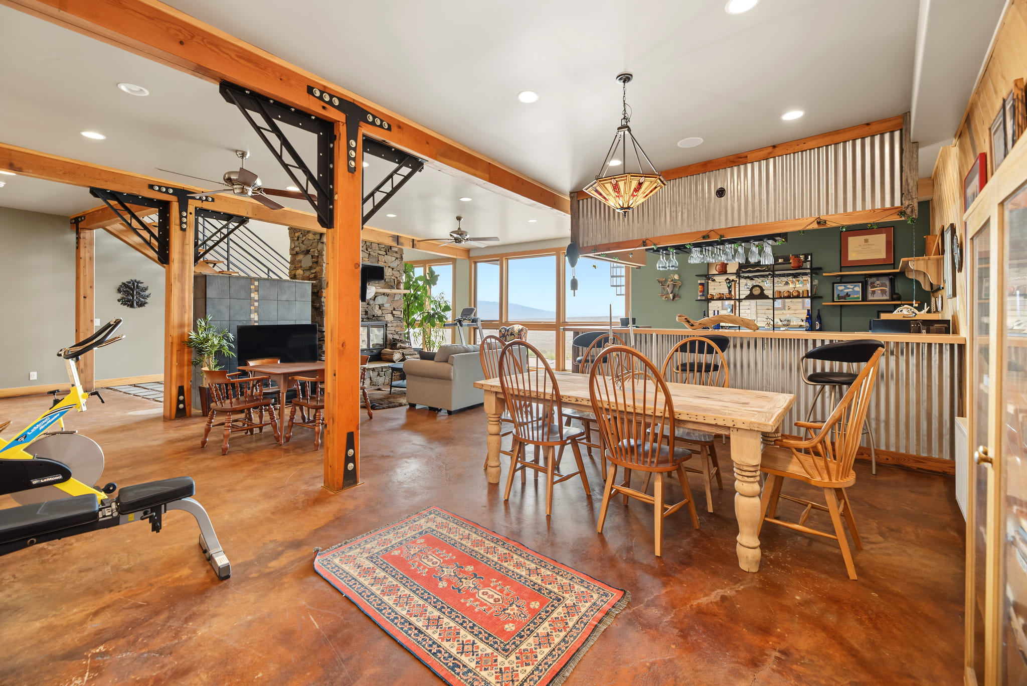 Open-concept room with wood beams, dining table, bar area, and exercise bike. Large windows offer natural light. The space has a rustic-industrial style with corrugated metal and wood accents. A red rug is on the polished floor.