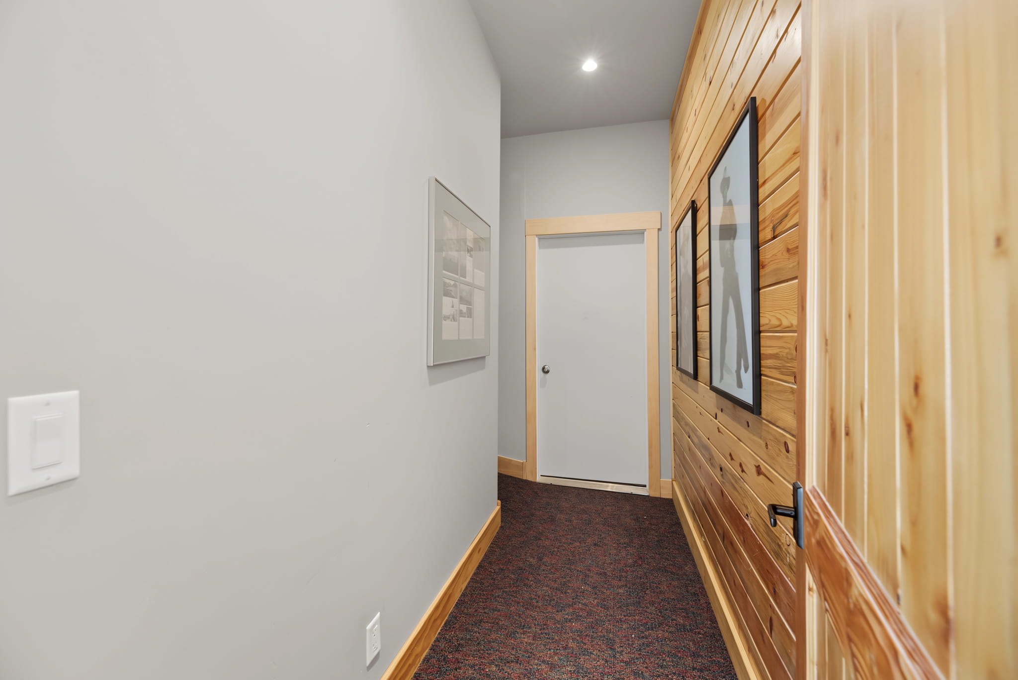 A hallway with light gray walls, one wall covered in wooden panels, framed art, a white door at the end, and dark red carpeting.