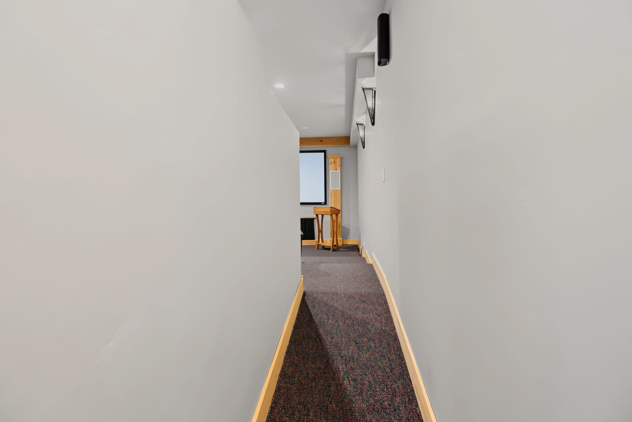 A narrow hallway with light gray walls, dark carpet, and wood trim leads to a room with a projector screen and a wooden lectern.