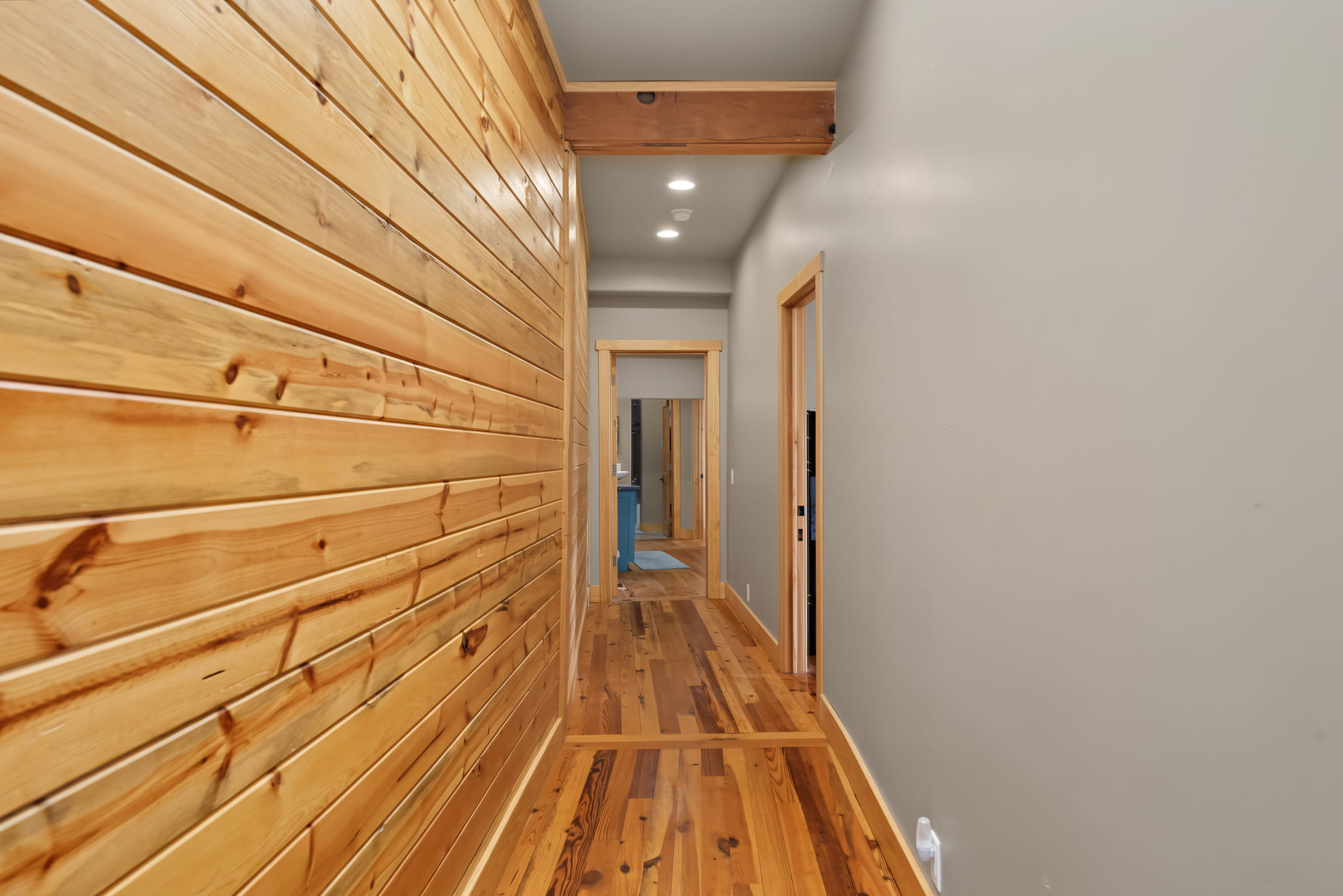 A hallway with wood paneling on one wall and hardwood flooring. The opposite wall is painted light gray, and there are doors along the hallway leading to other rooms, with ceiling lights visible above.