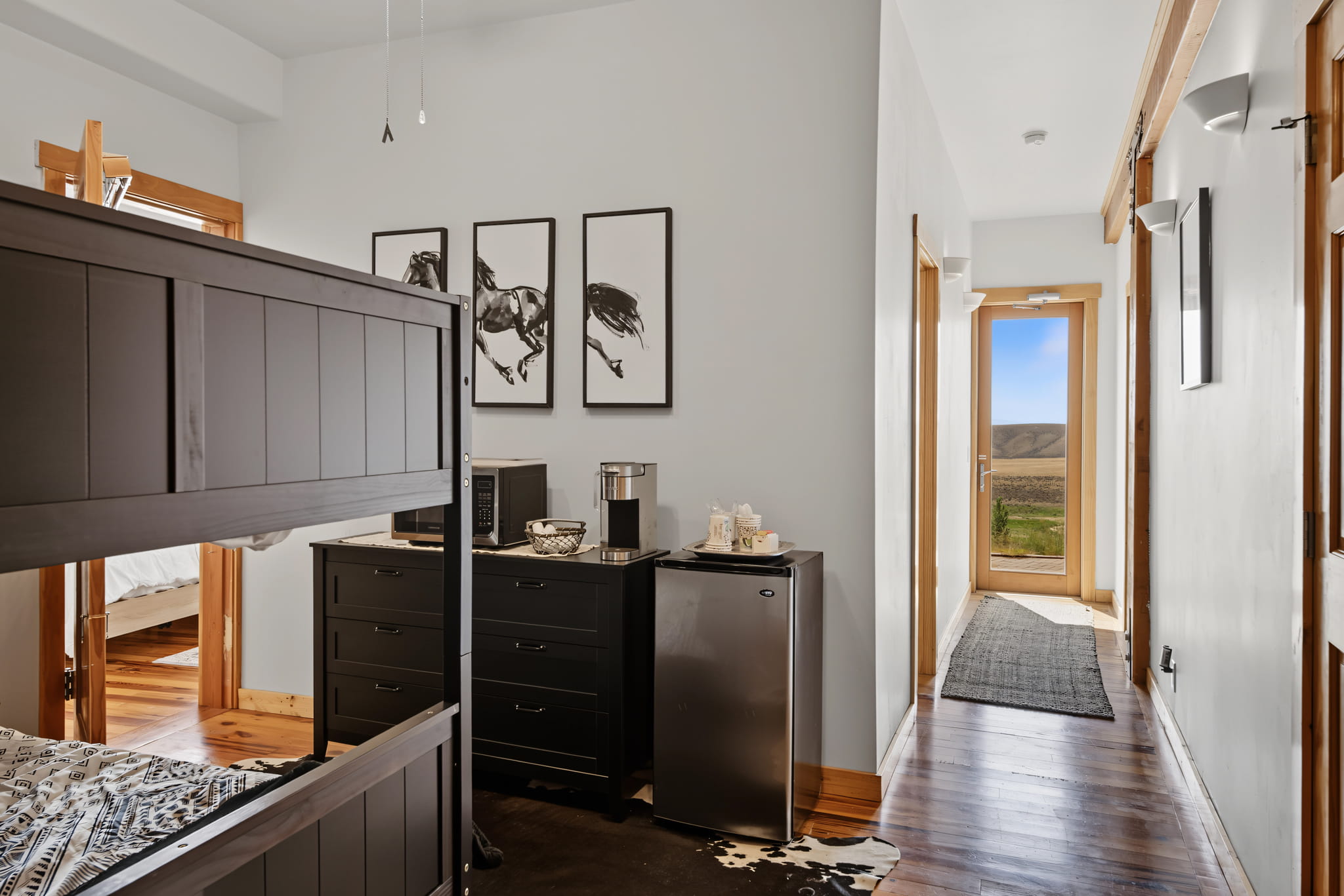 A bunk bed, black dresser with a microwave, coffee maker, and mini fridge, art of horses on the wall, wood floors, and a hallway leading to a door with a scenic outdoor view.