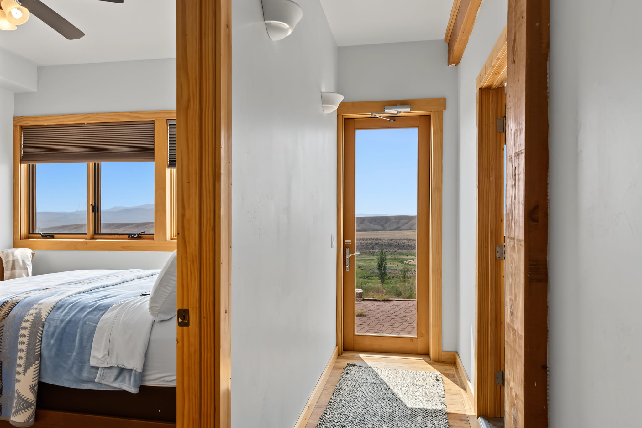 A hallway with wood trim leads to a glass door opening to a patio with scenic mountain views; a bedroom with a bed and window is visible on the left.