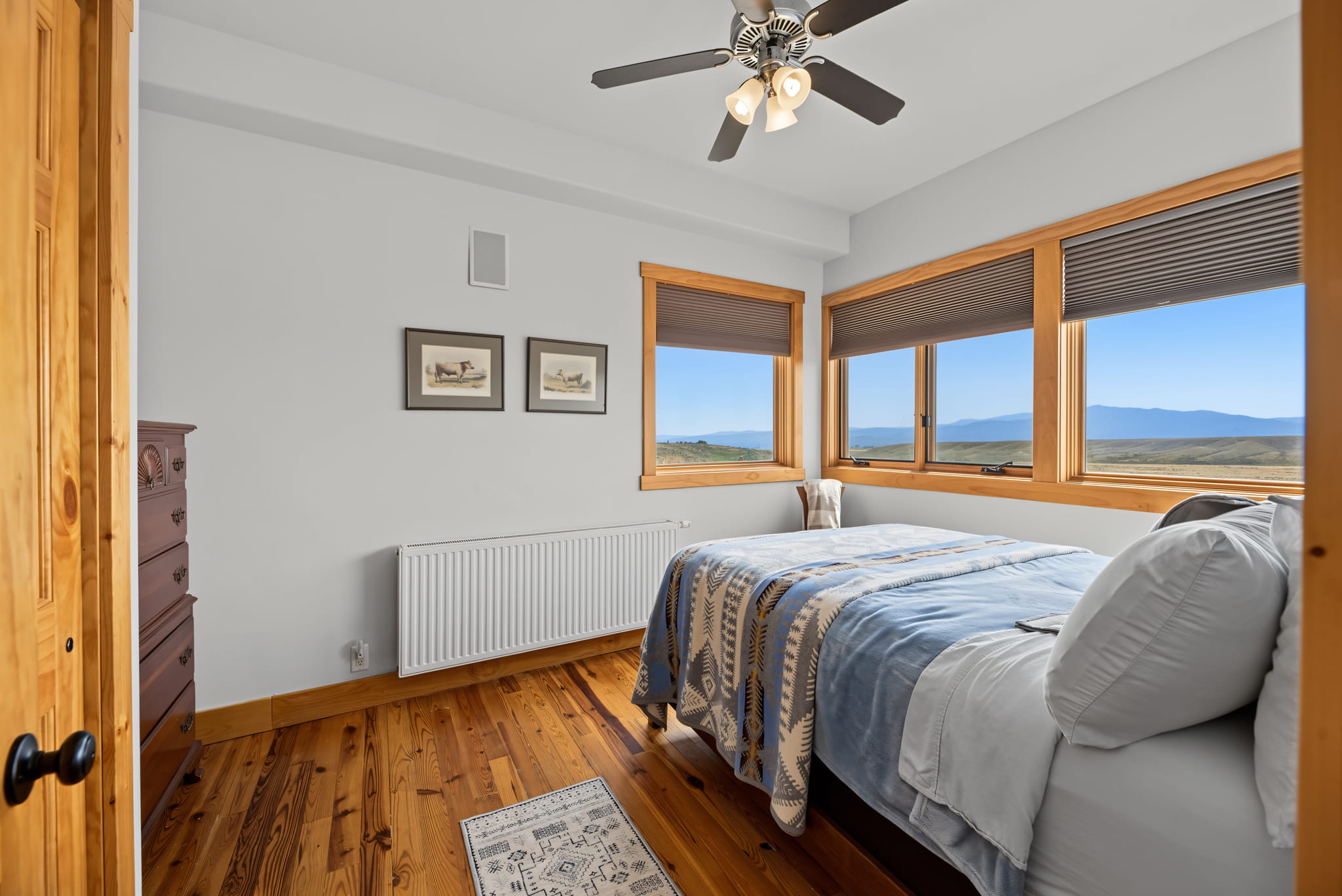 A cozy bedroom with wooden floors, a bed with blue bedding, a ceiling fan, a dresser, two framed pictures, and large windows with mountain views.