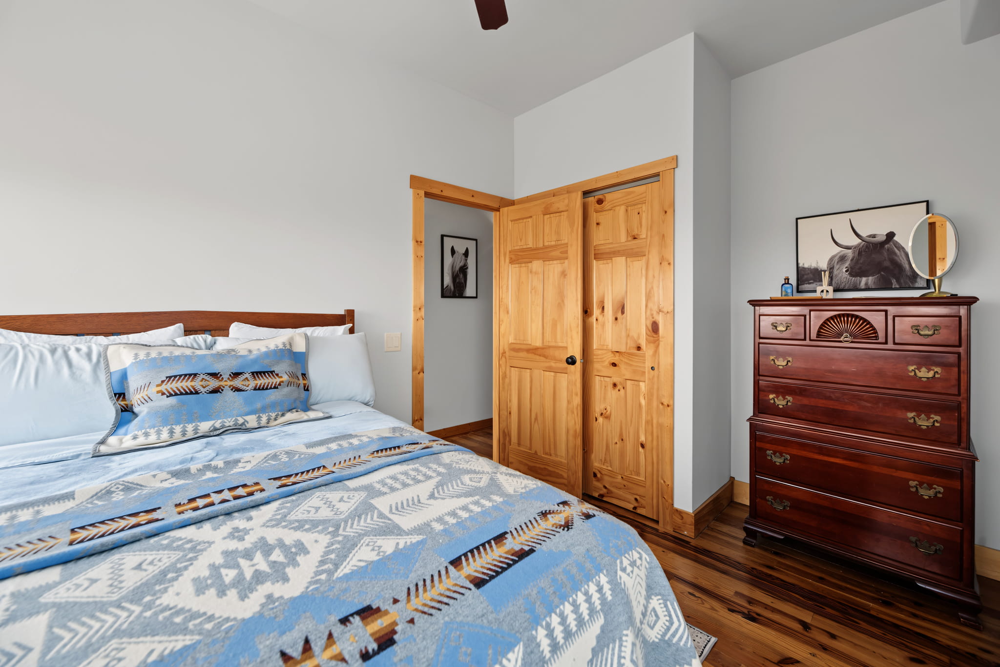 A cozy bedroom with a patterned blue and white bedspread, wooden floors, a light wood door, a dark wood dresser, and animal art on the white walls.