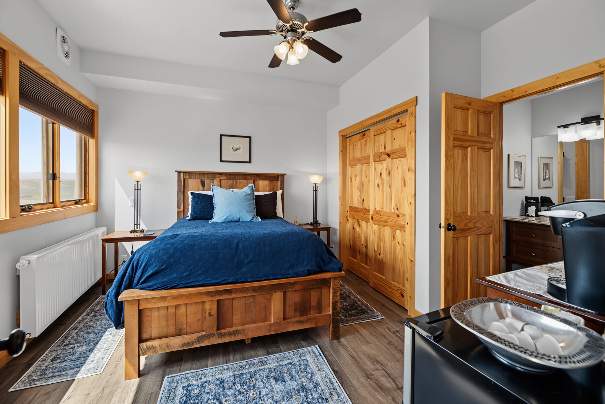 Cozy bedroom with wooden bed, blue bedding, two bedside tables, lamps, ceiling fan, and closet. Window lets in natural light. Mini fridge and coffee maker are in the foreground. Rug adds warmth to wooden floor.