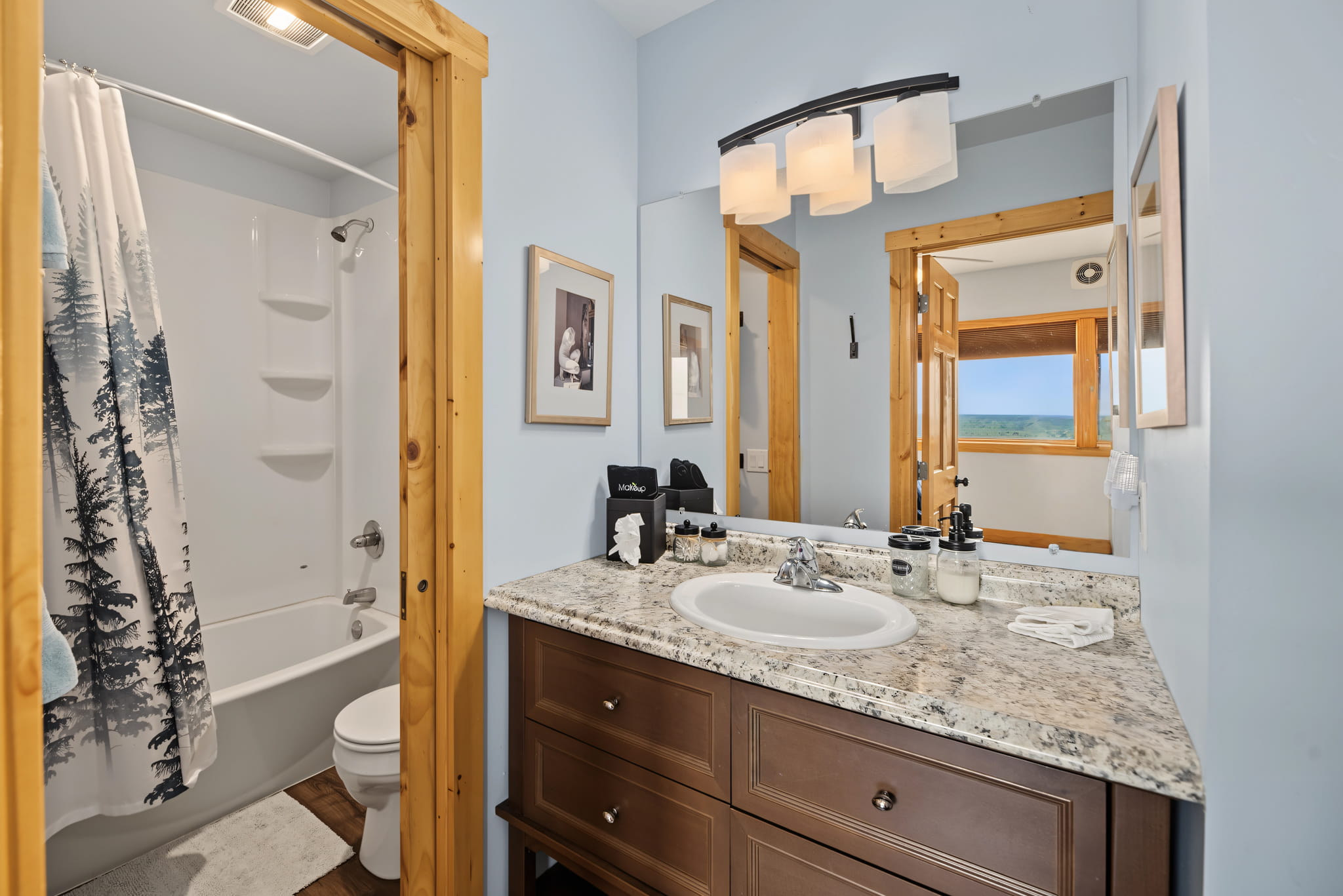 A bathroom with a granite countertop, sink, wooden vanity, wall mirror, and a bathtub with a tree-patterned shower curtain. A window in the hallway offers a scenic view.