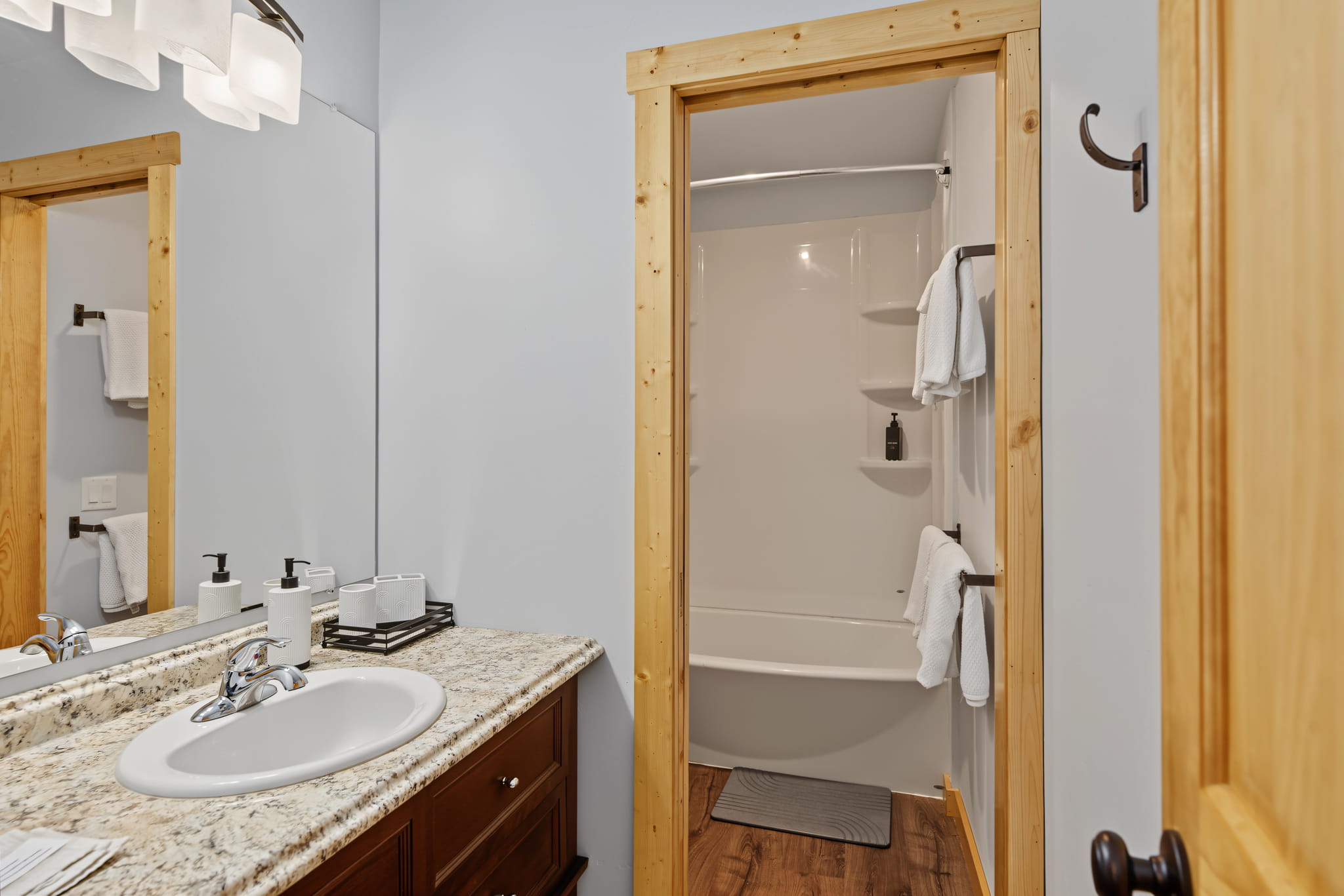 Modern bathroom with a granite countertop, sink, and mirror. The doorway leads to a white bathtub with shelves and towels. Light wood trim frames the door and mirror, with white walls and wood flooring throughout.