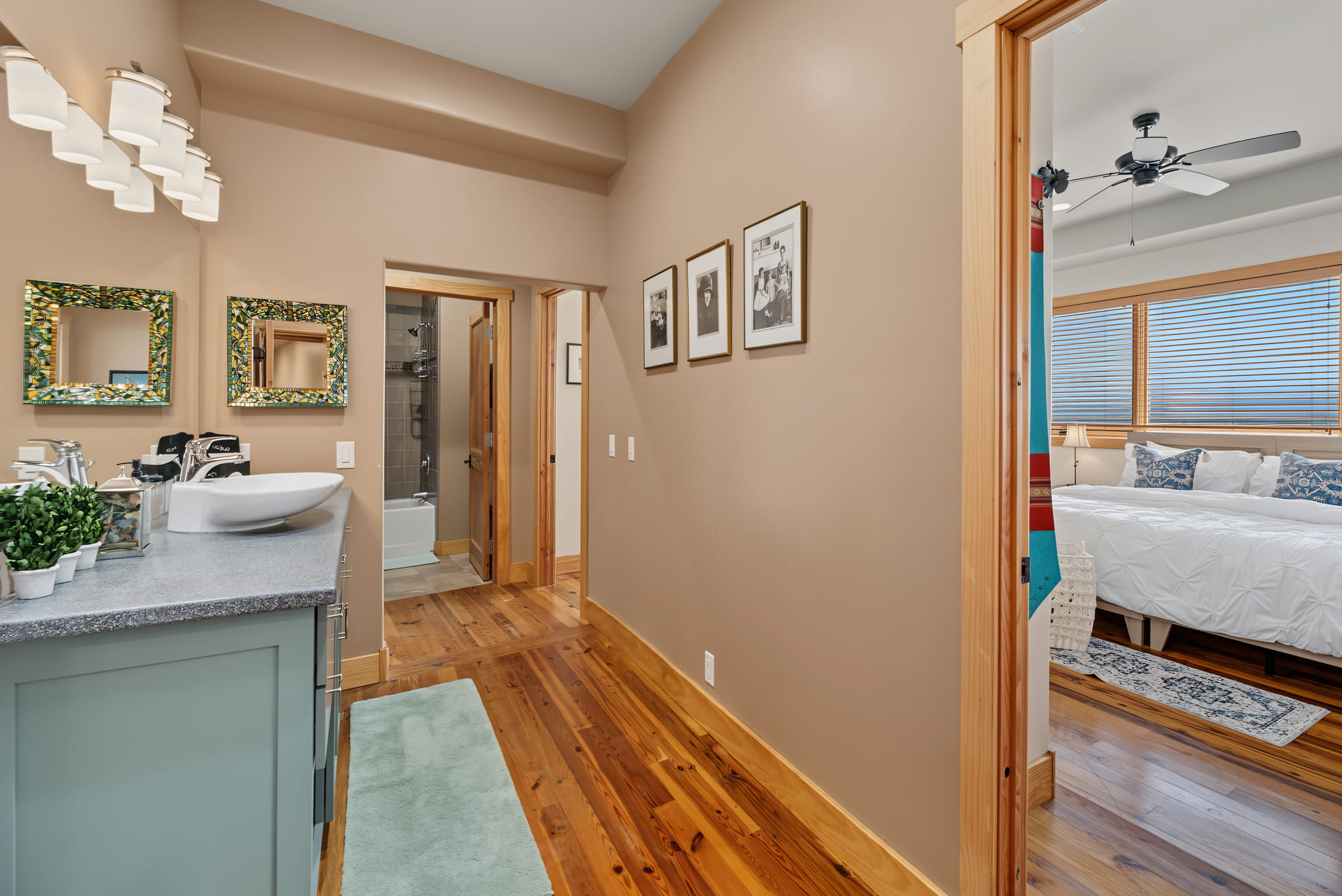 A hallway with wood floors leads to a bathroom on the left with a vanity and green mirrors, and a bedroom on the right with a white bed and large windows. Framed photos hang on the beige hallway wall.