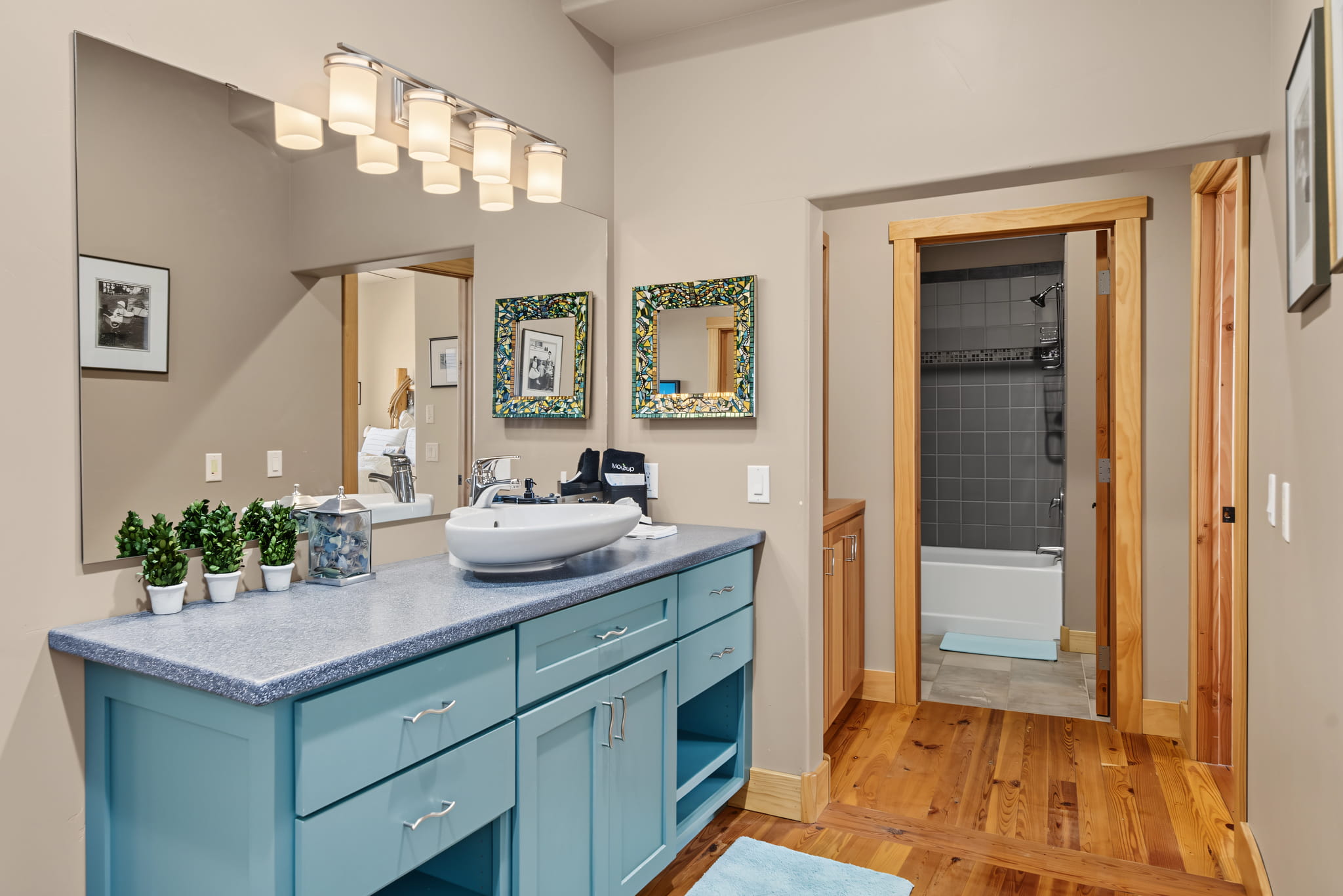 A bathroom with a blue vanity, potted plants, a vessel sink, two mirrors, and a wall-mounted light fixture. A bathtub and gray tiled shower are visible through an open doorway.