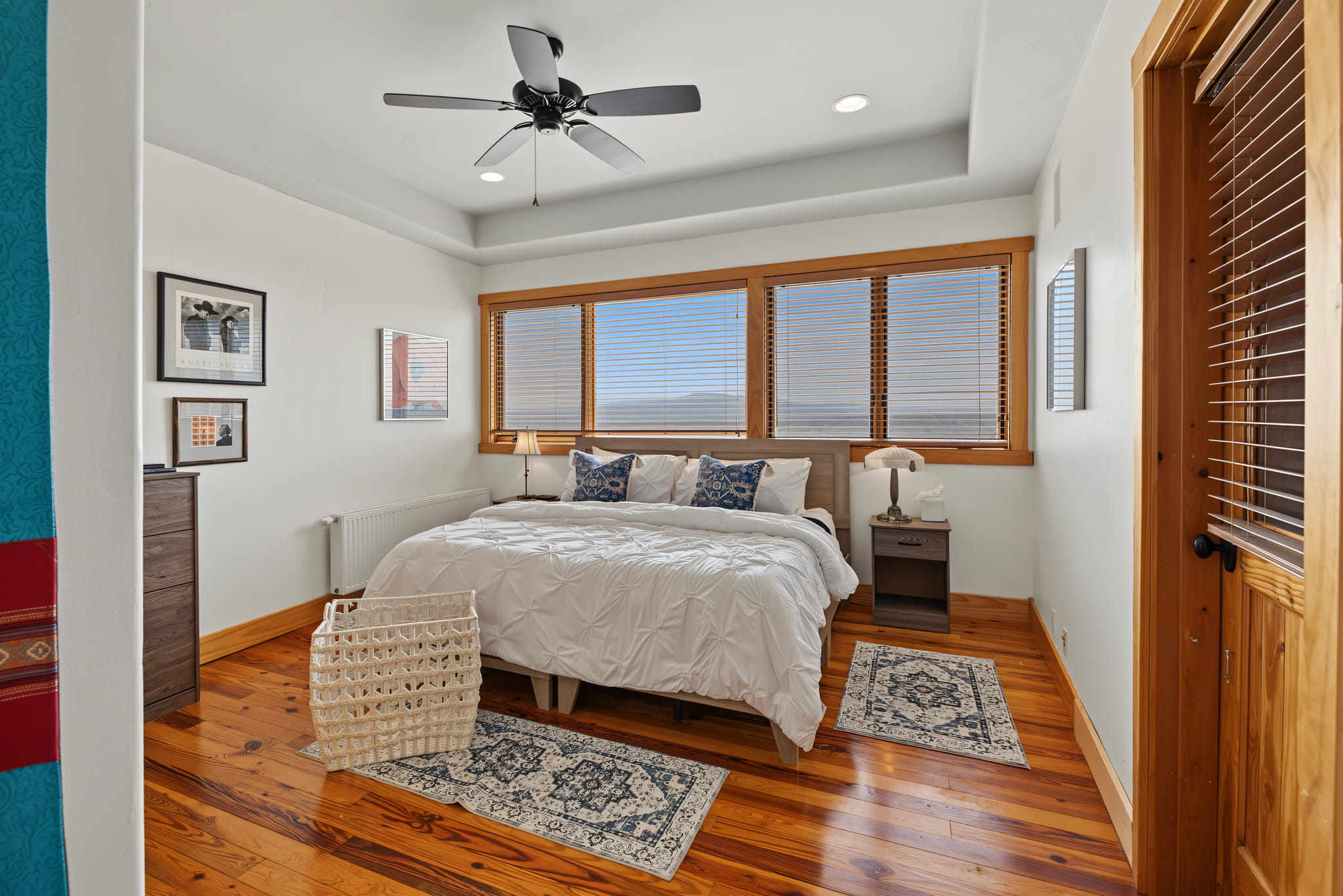 Bright bedroom with wooden floors, a ceiling fan, large windows with blinds, a neatly made bed with white linens, two bedside tables, wall art, two rugs, and a white laundry basket.