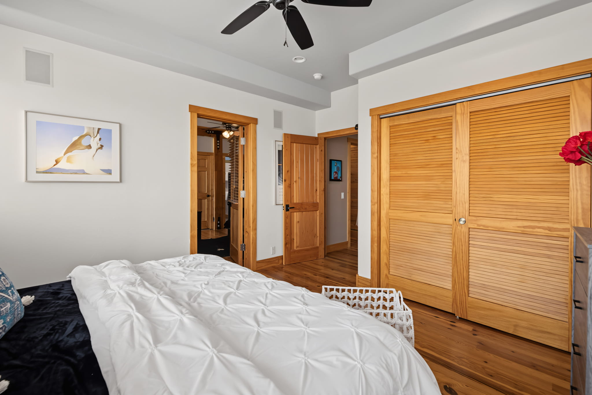 A cozy bedroom with white bedding, wooden floors, a ceiling fan, wooden doors, a framed art piece on the wall, and a white laundry basket at the foot of the bed.