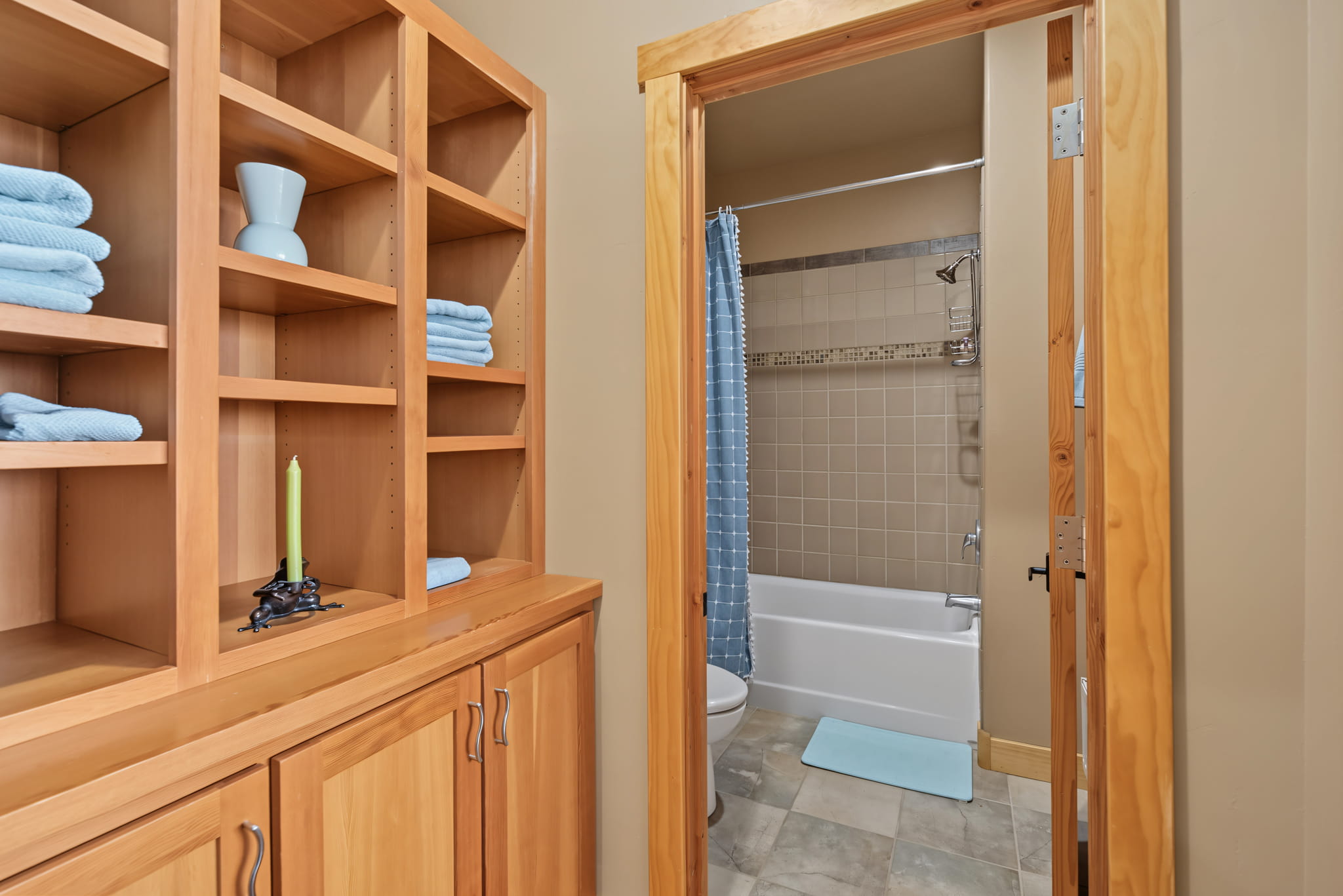 Open wooden shelves with folded blue towels and a vase, next to a doorway leading to a bathroom with a blue shower curtain, a white tub, and a blue bathmat on a tiled floor.