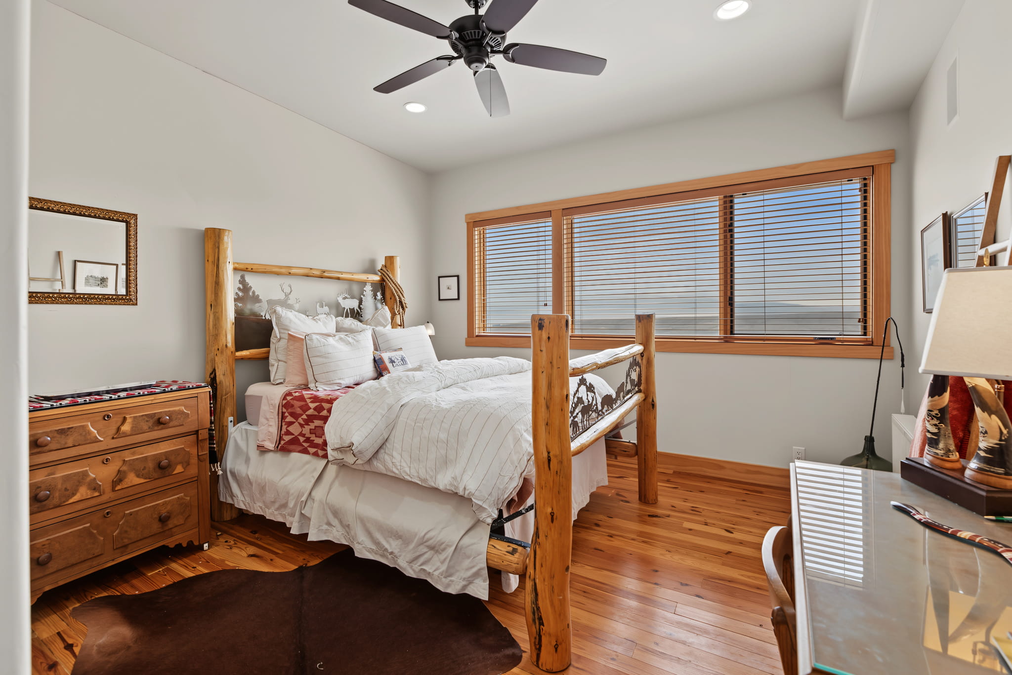 A cozy bedroom with a rustic wooden bed, white bedding, a dresser, desk, lamps, cowhide rug, and large windows with blinds letting in natural light. Ceiling fan and wooden floor add warmth to the room.