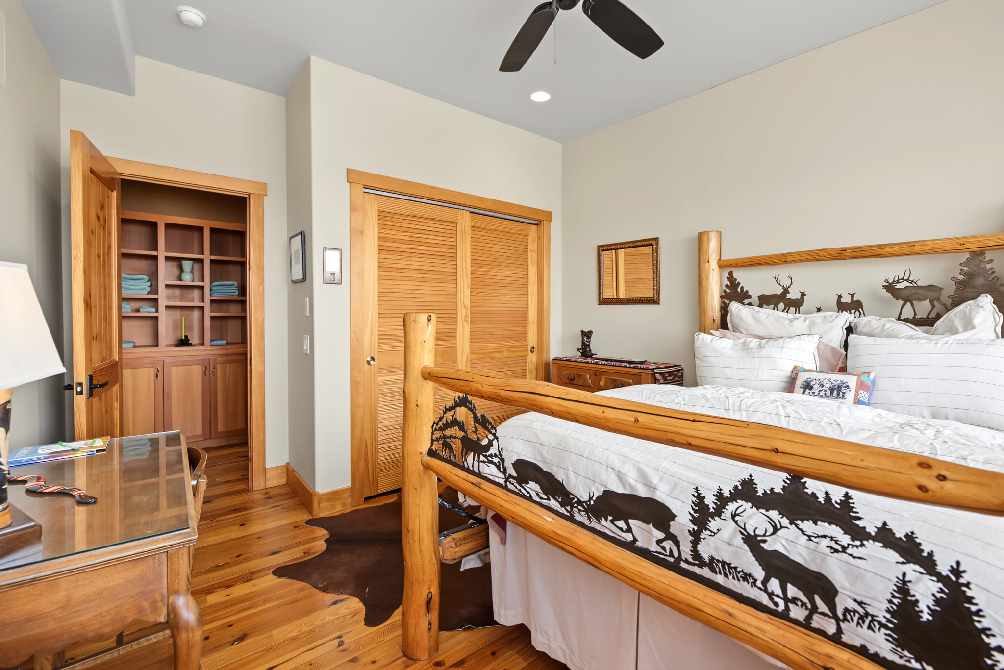 A cozy bedroom with wood furniture, rustic wildlife-themed bedding, wood floors, a desk with a lamp, a ceiling fan, a small closet, and built-in shelves with folded towels.