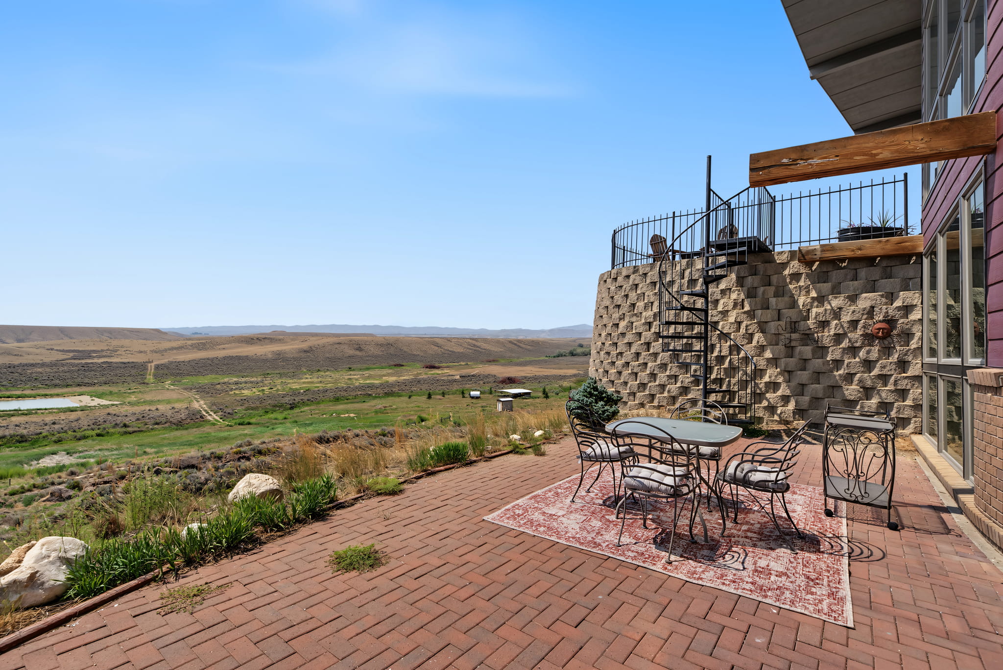 Outdoor patio with a table and chairs on a rug, overlooking a vast, hilly landscape. Spiral staircase leads to an upper deck on a stone wall. Blue sky and open countryside in the background.