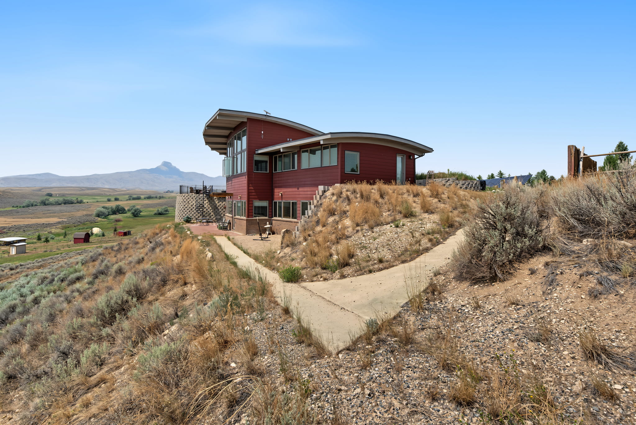 A modern red house with large windows sits on a dry, sloped landscape with a concrete path. Sparse shrubs cover the ground, and mountains are visible in the background under a clear blue sky.