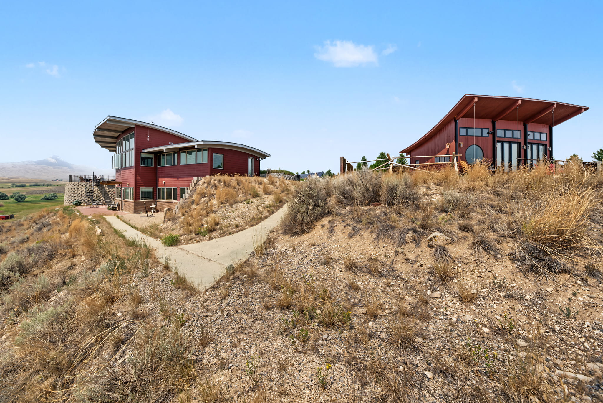 Two modern red buildings with large windows sit on a dry, rocky hillside with sparse grass and plants. A concrete pathway leads between them under a clear blue sky. Mountains are visible in the distance.