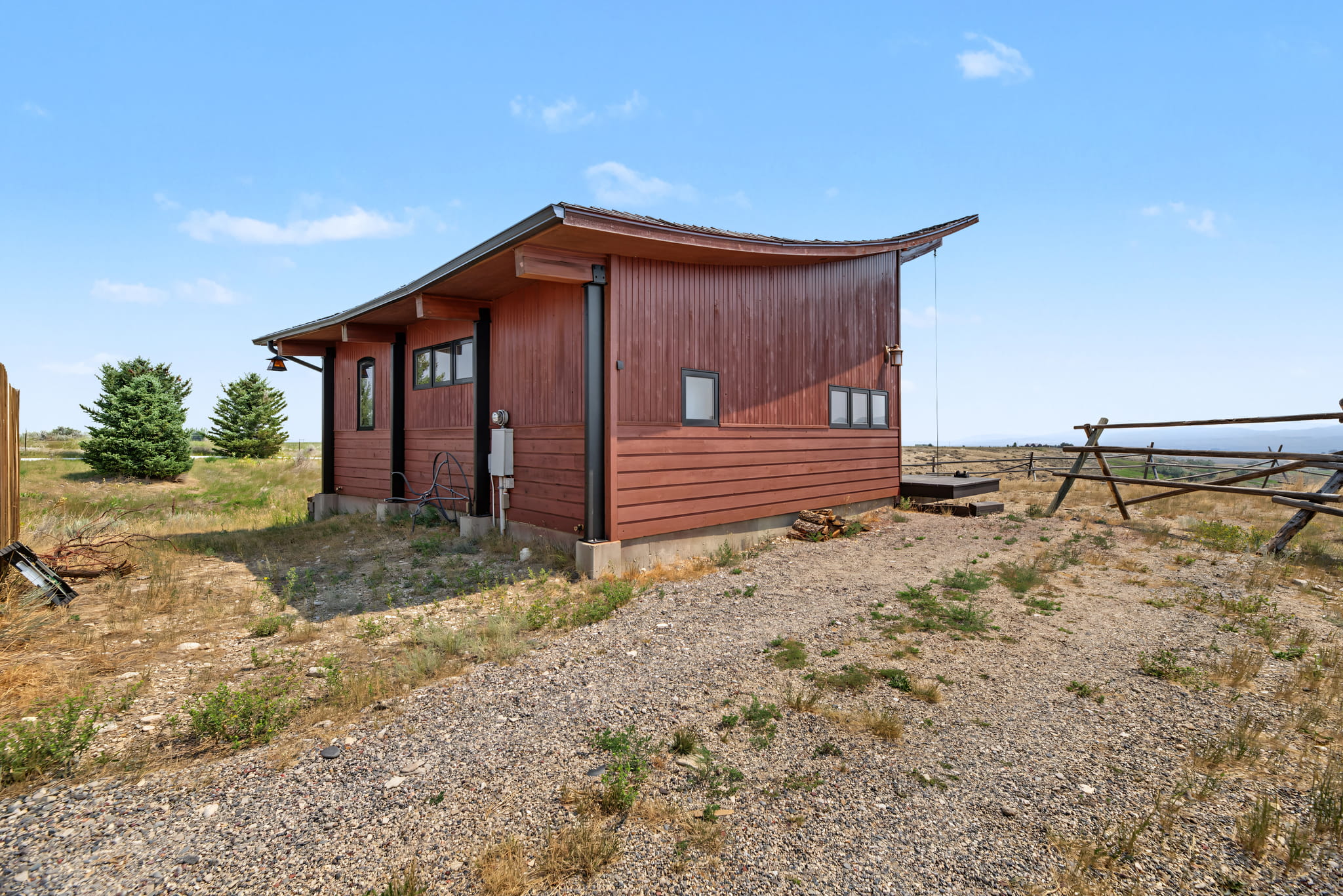 A small modern wooden cabin with a sloped roof sits on a gravel lot surrounded by grass and open land, under a blue sky with scattered clouds.