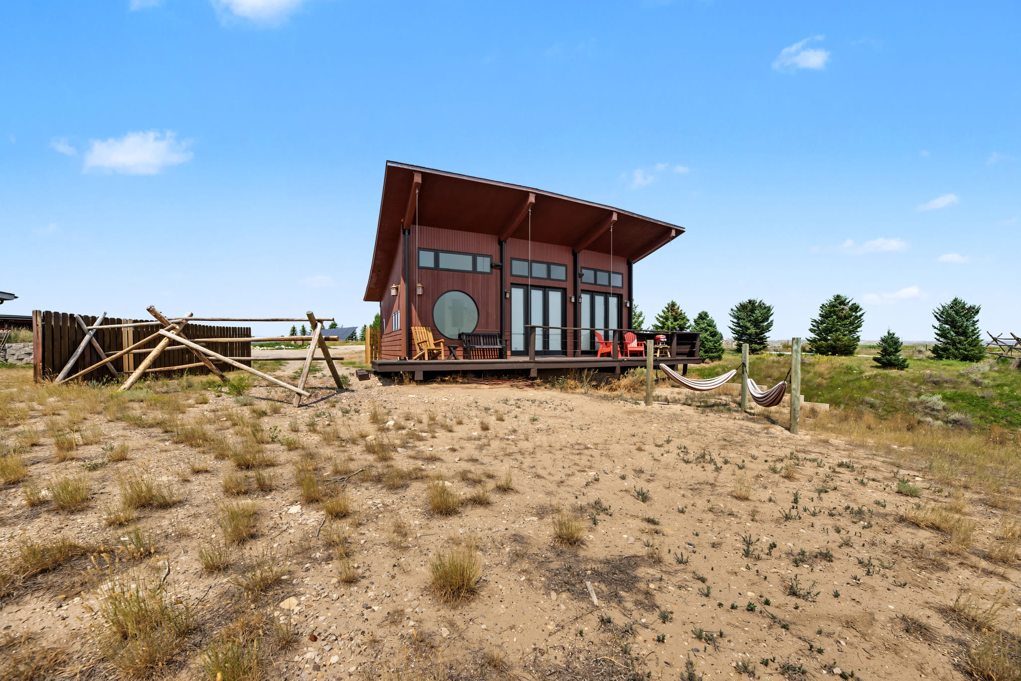 A modern cabin with large windows and a slanted roof sits on a wooden deck in a dry, grassy landscape. Hammocks hang between posts, and Adirondack chairs face the open view. Pine trees and blue sky are in the background.