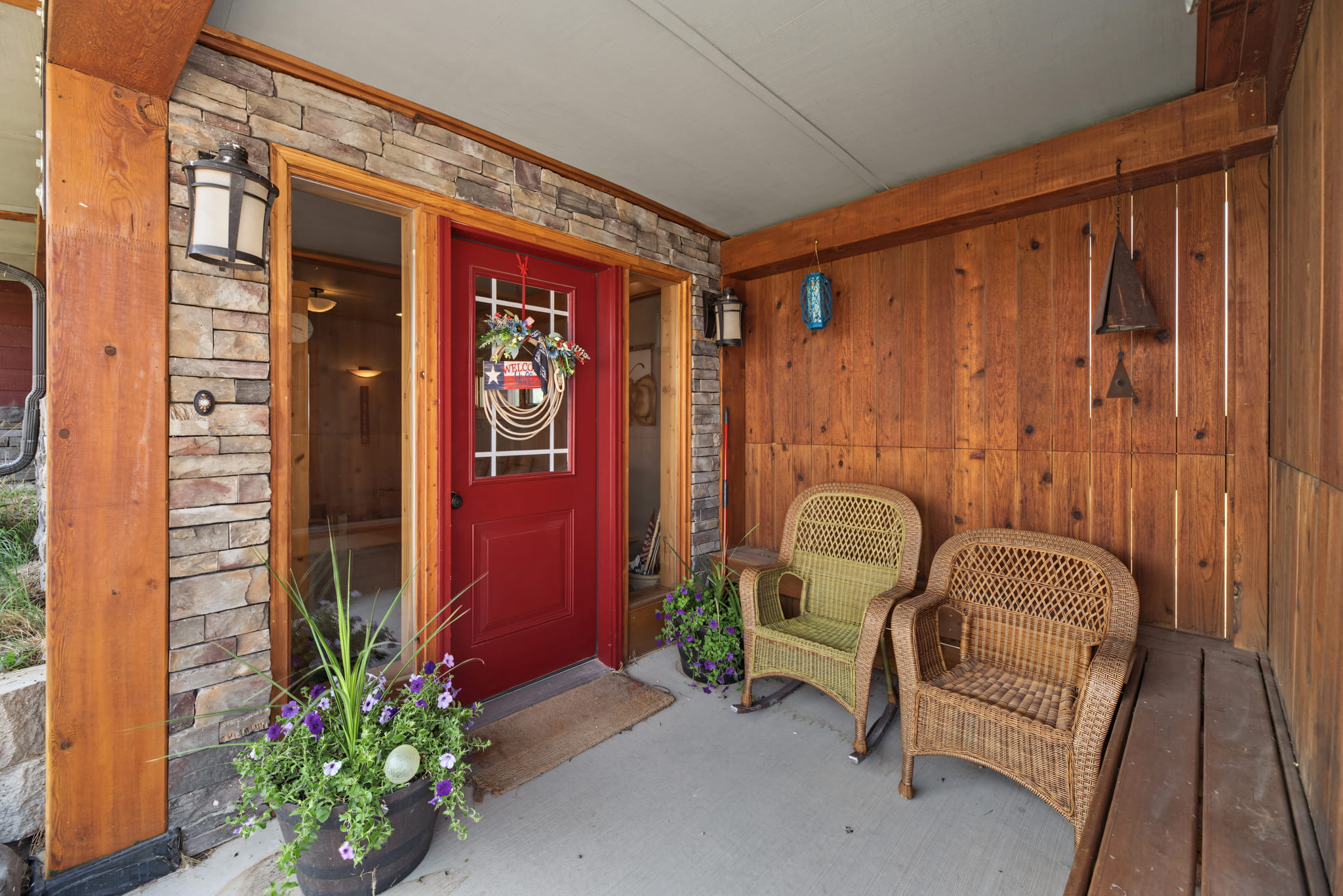 A cozy porch with a red door, a welcome wreath, two wicker chairs, potted flowers, wood paneling, and stone wall accents.