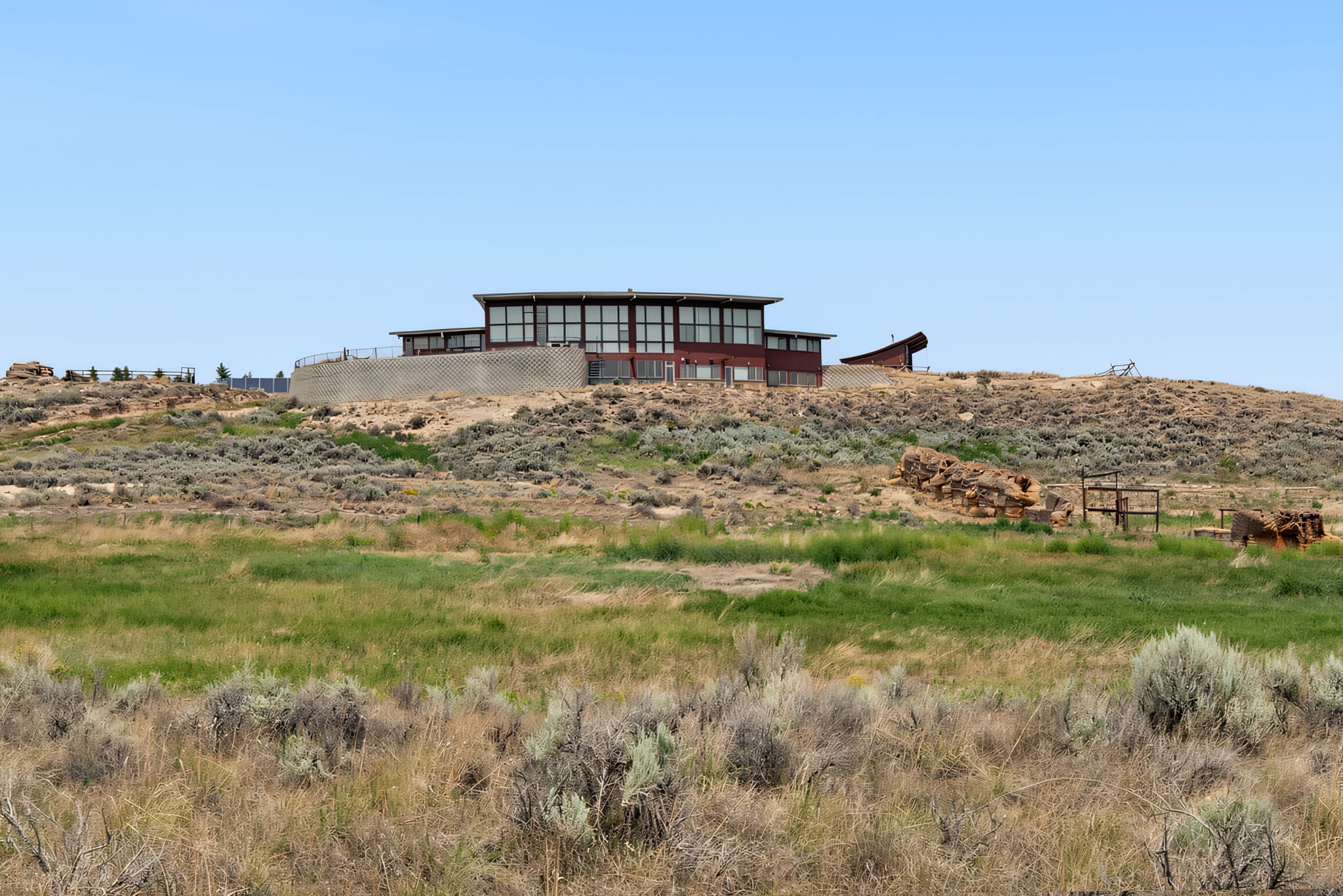 A modern, circular building with large windows sits atop a dry, grassy hill under a clear blue sky. The landscape features sparse shrubs and patches of green vegetation.