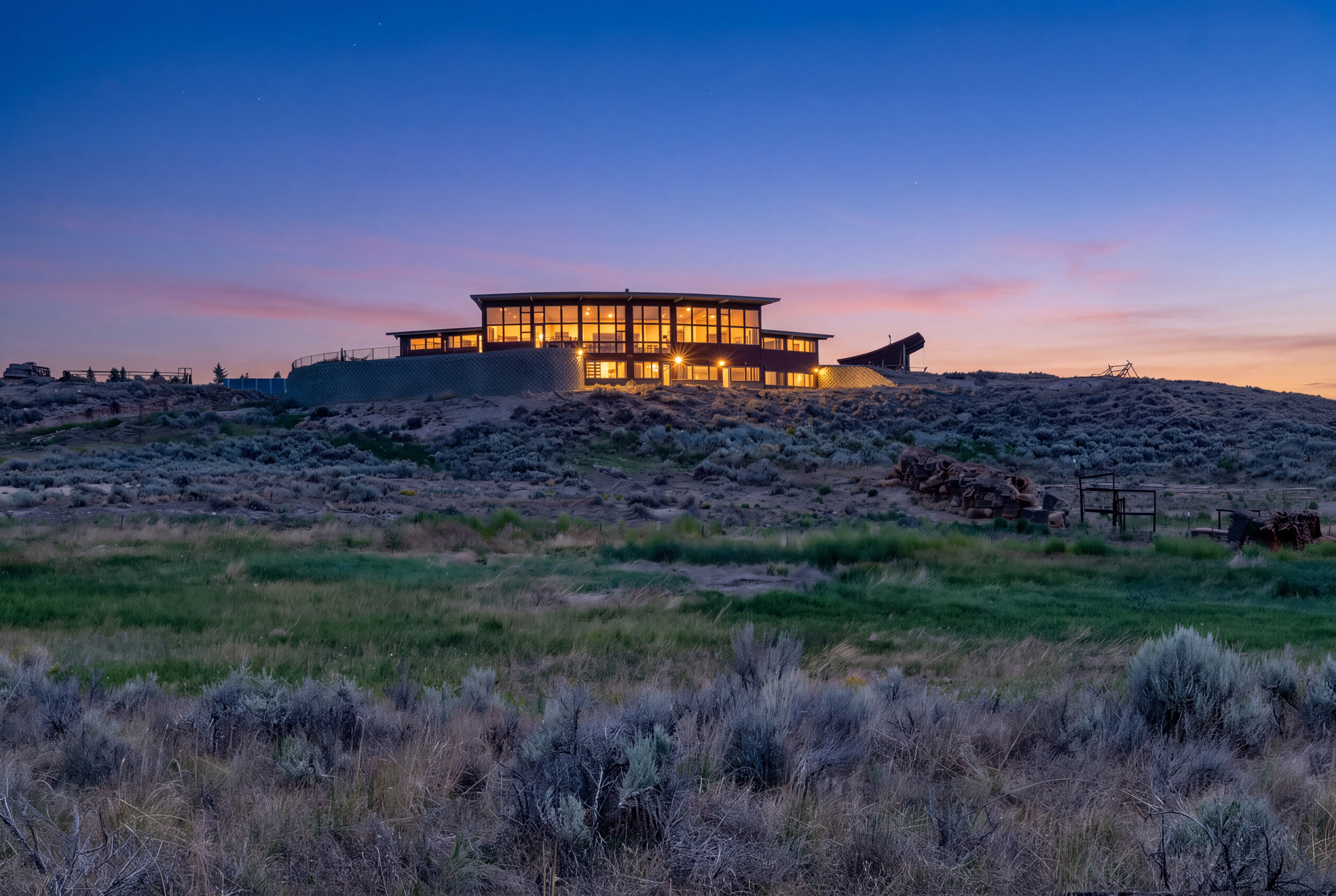 A modern building with glowing windows sits atop a hill at sunset, surrounded by dry grass and shrubs under a clear twilight sky.